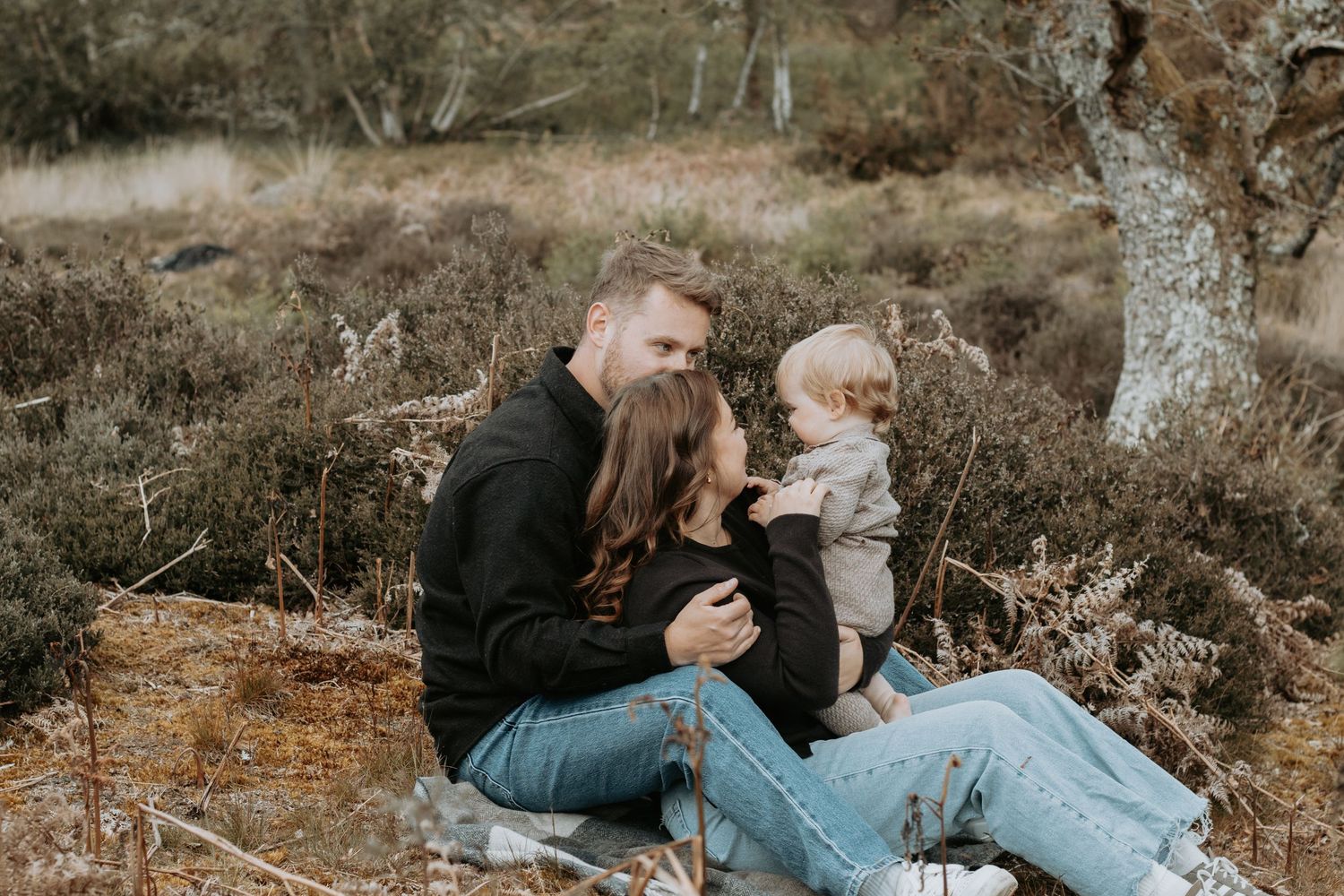A young family sitting outdoors in a natural setting, parents lovingly gazing at their baby amid wild shrubs.