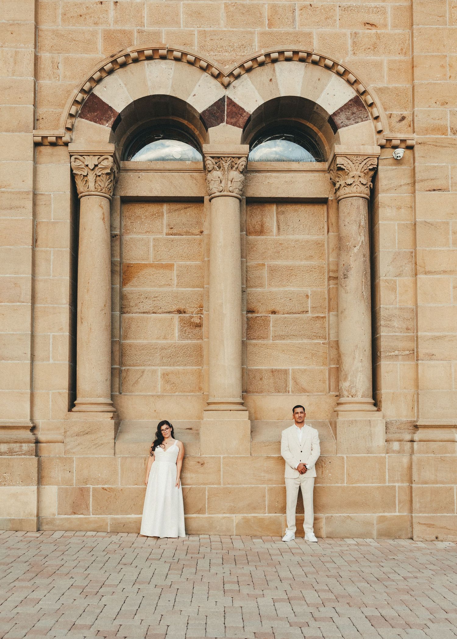 Couple in white attire posing before ornate sandstone arched windows of a historic building.