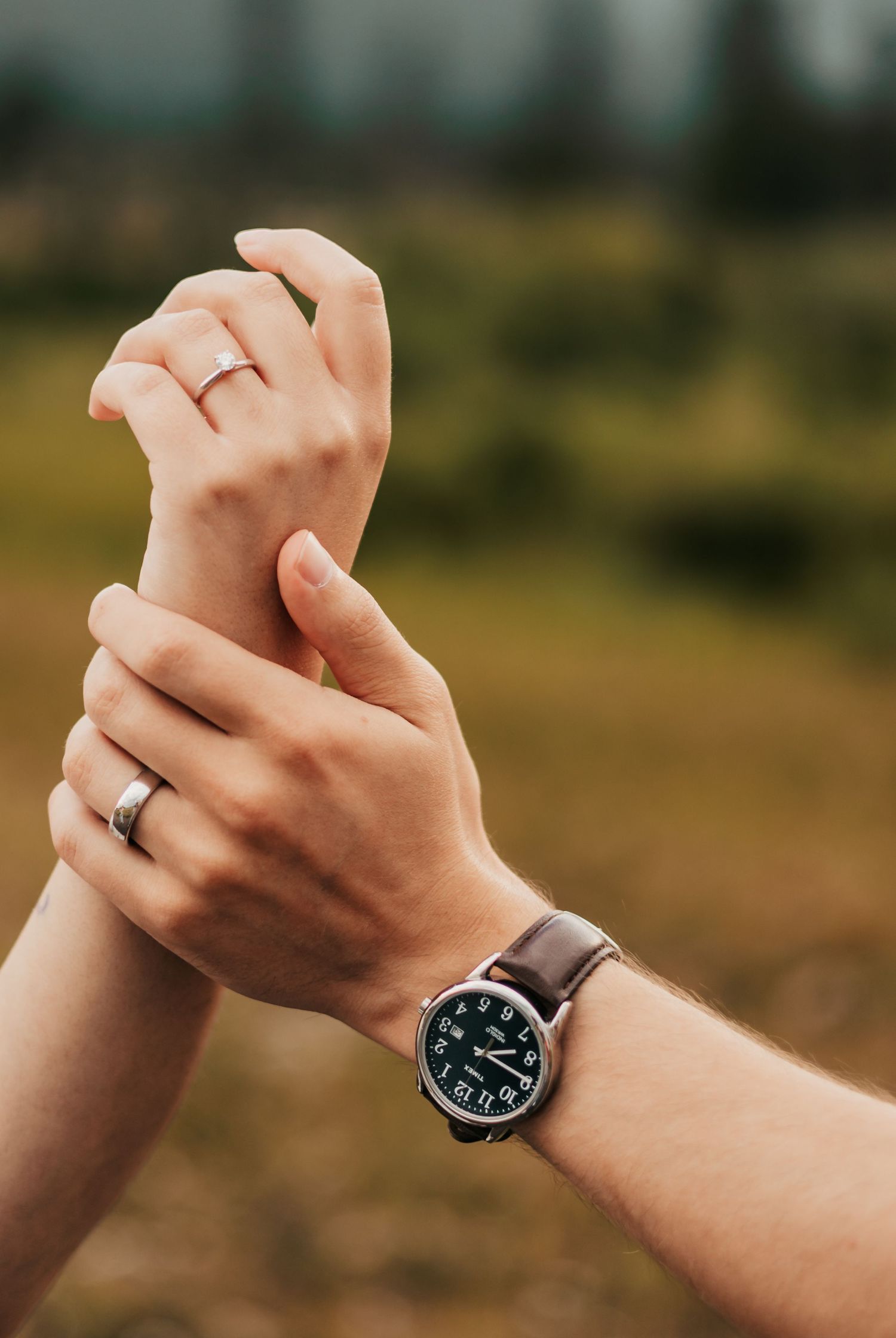 Two hands reaching toward each other, one with a leather bracelet and one with a ring, against a blurred green background.