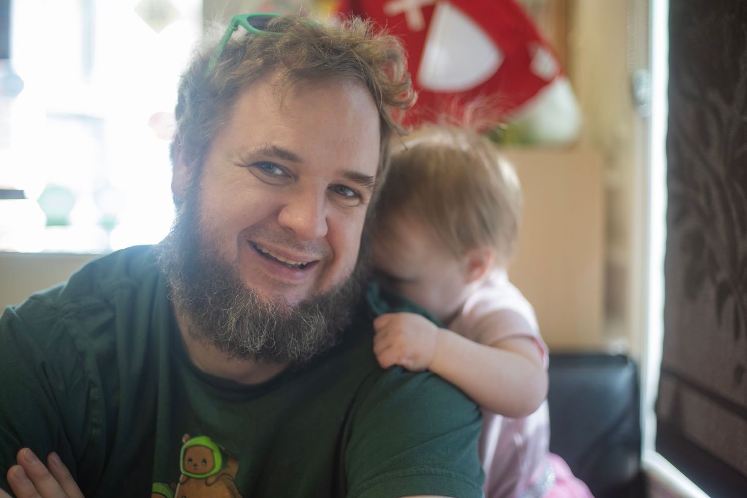 Smiling bearded man in green shirt holds a baby resting on his shoulder in a cozy indoor setting.