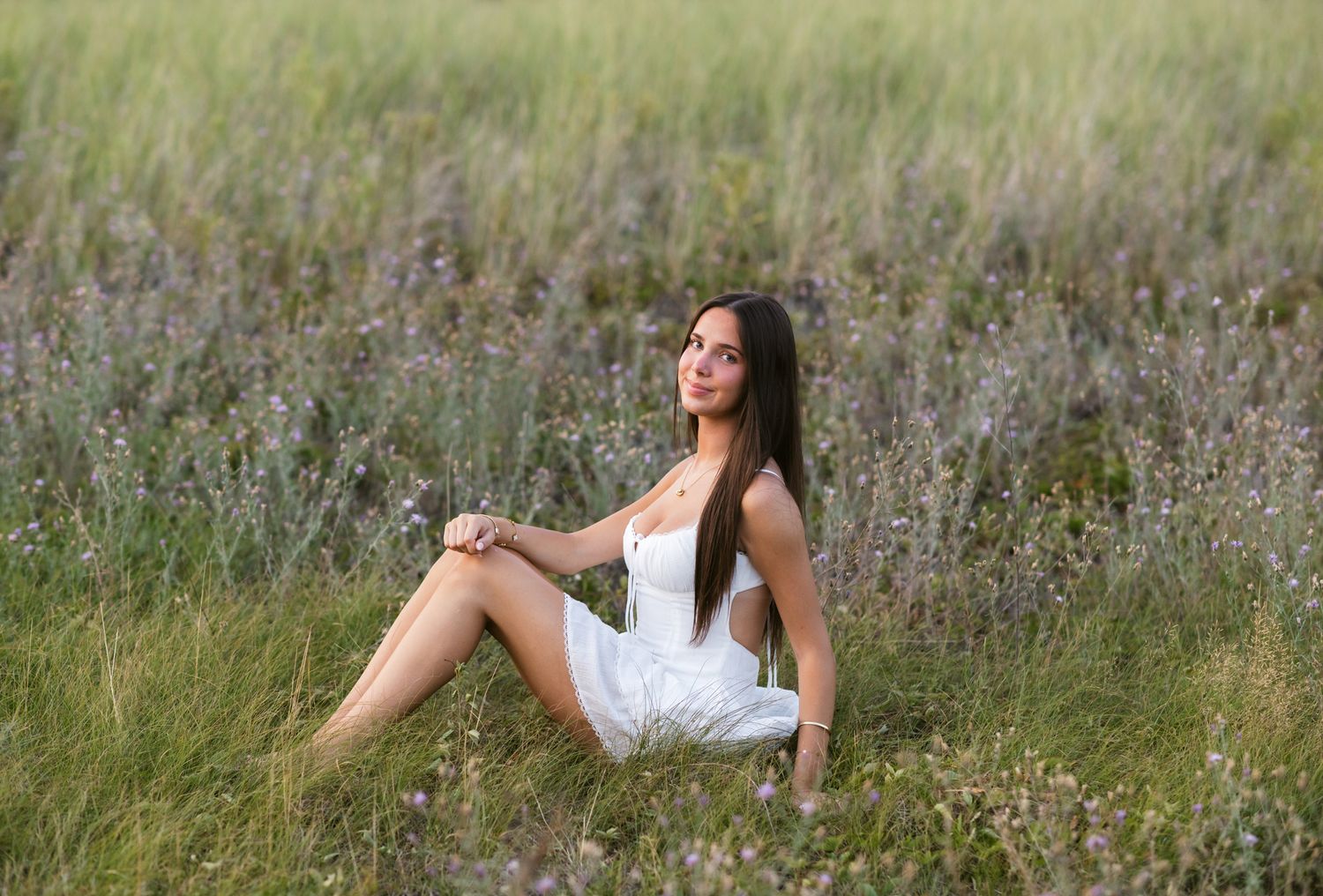 Teen girl sits in a field of clover wearing a white dress for senior photos at Salisbury State Reservation. Seacoast NH senior photographer.