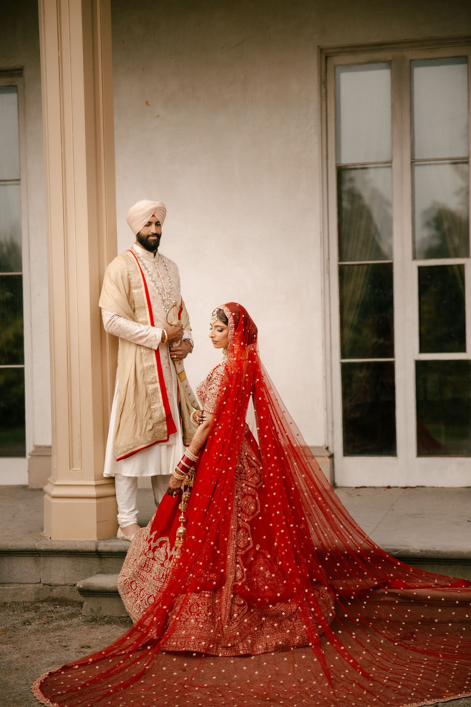 Indian bride in red lehenga and groom in cream sherwani stand apart on green lawn with trees backdrop.