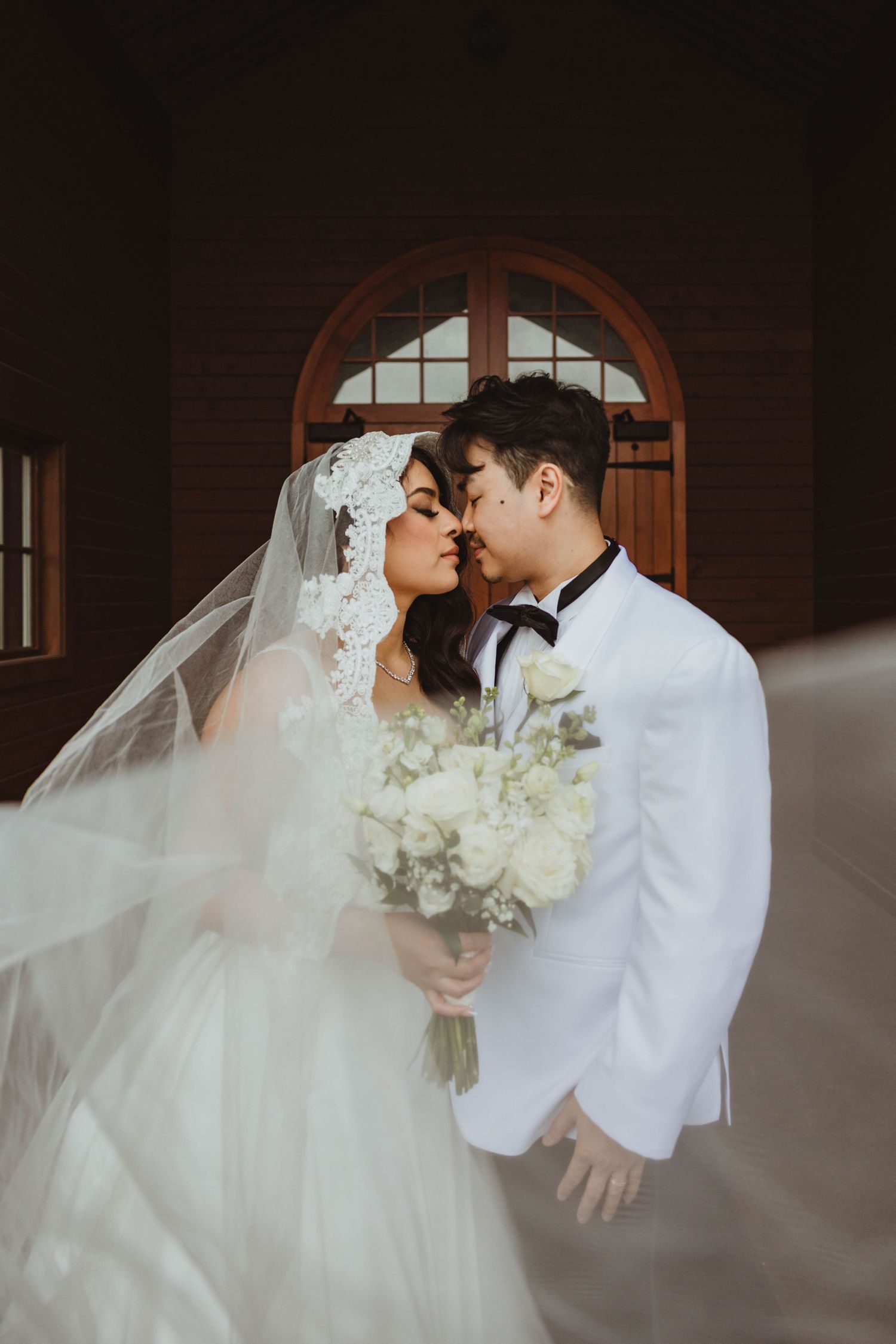 A bride and groom share a romantic kiss outside a wooden chapel, her veil flowing as she holds a white floral bouquet.