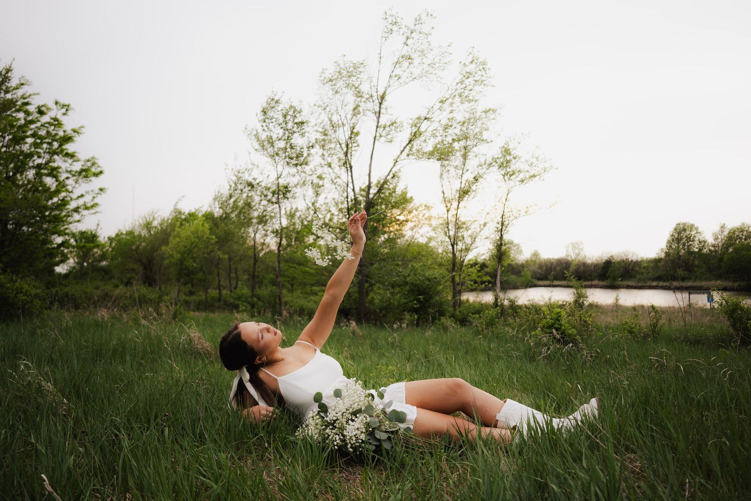 A young woman in a white dress lying in green grass holding a wildflower bouquet reaching skyward.