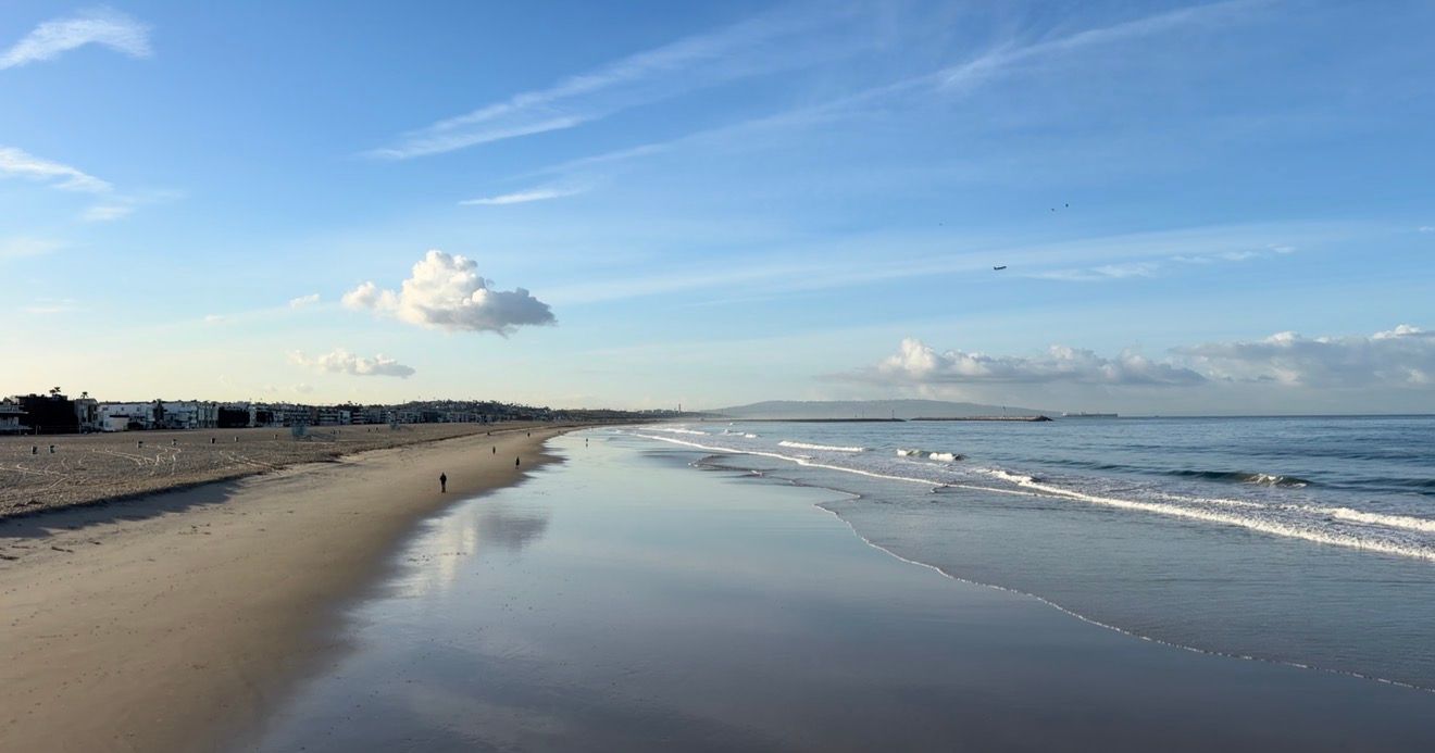 Serene beach shoreline with wet sand reflections, gentle waves, and a vast blue sky with scattered clouds on a calm day.