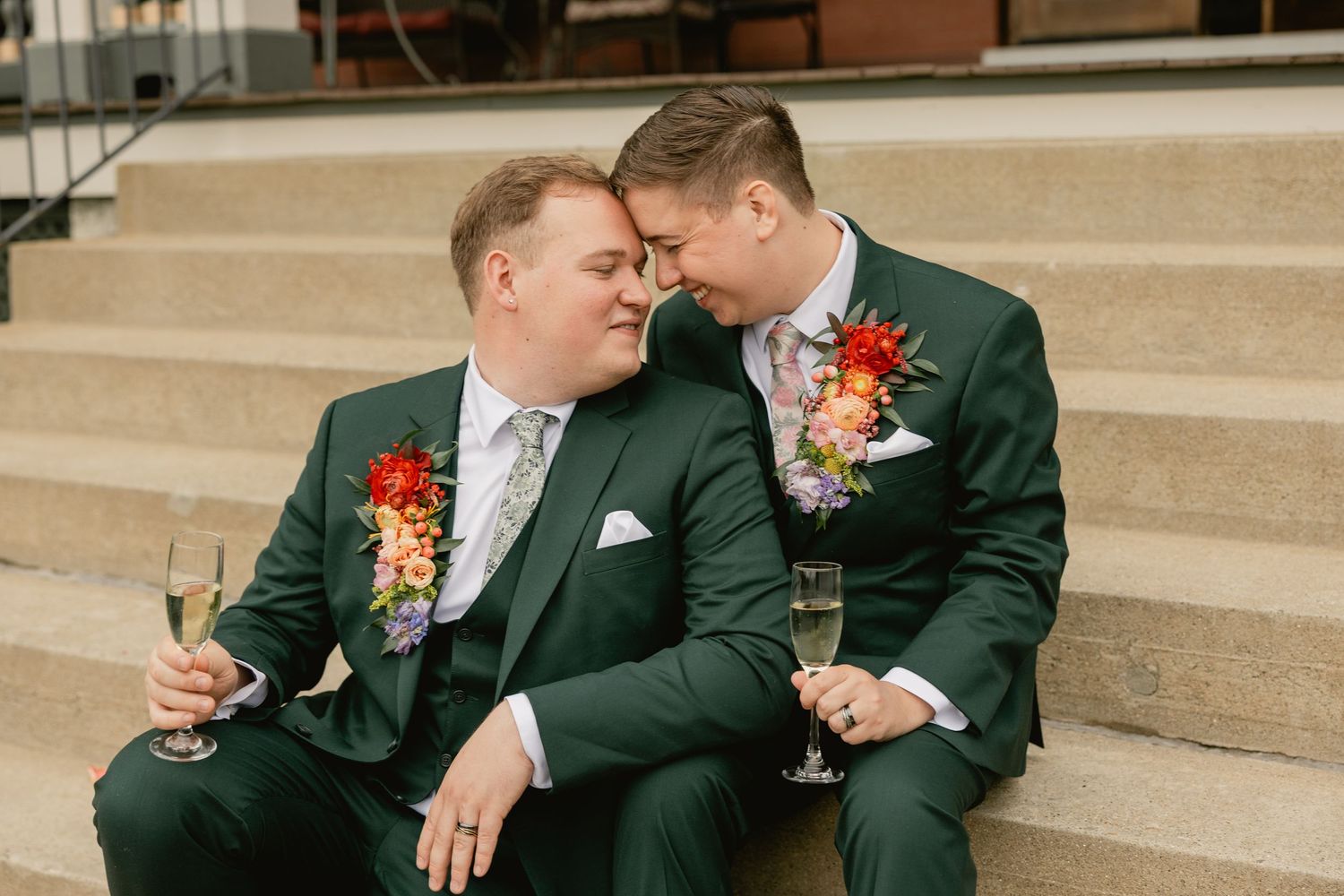 A couple in dark green attire shares an intimate moment on stone steps during a wedding celebration.