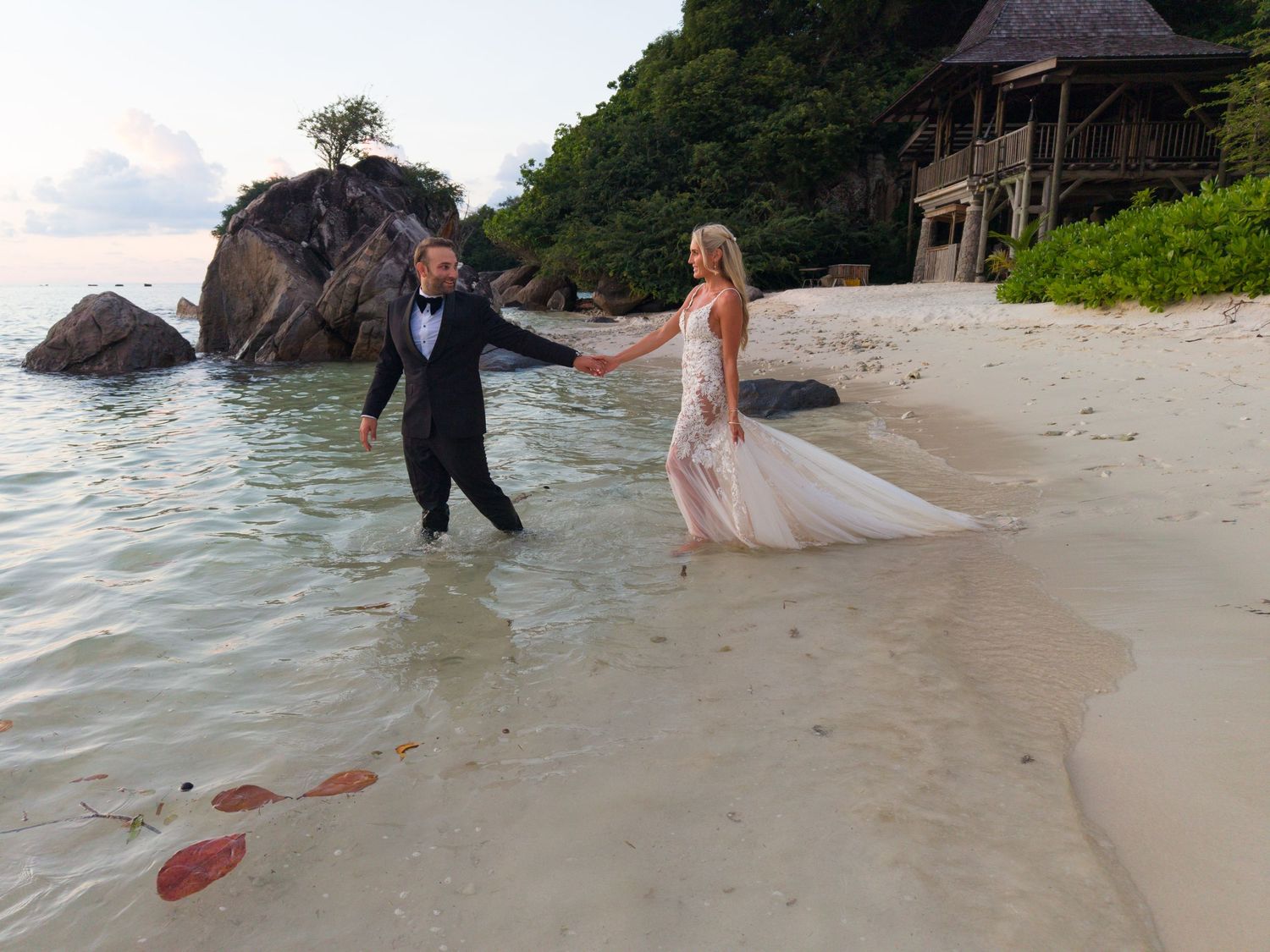 Groom lifting bride in white dress and veil on a tropical sandy beach during a romantic wedding photo shoot.