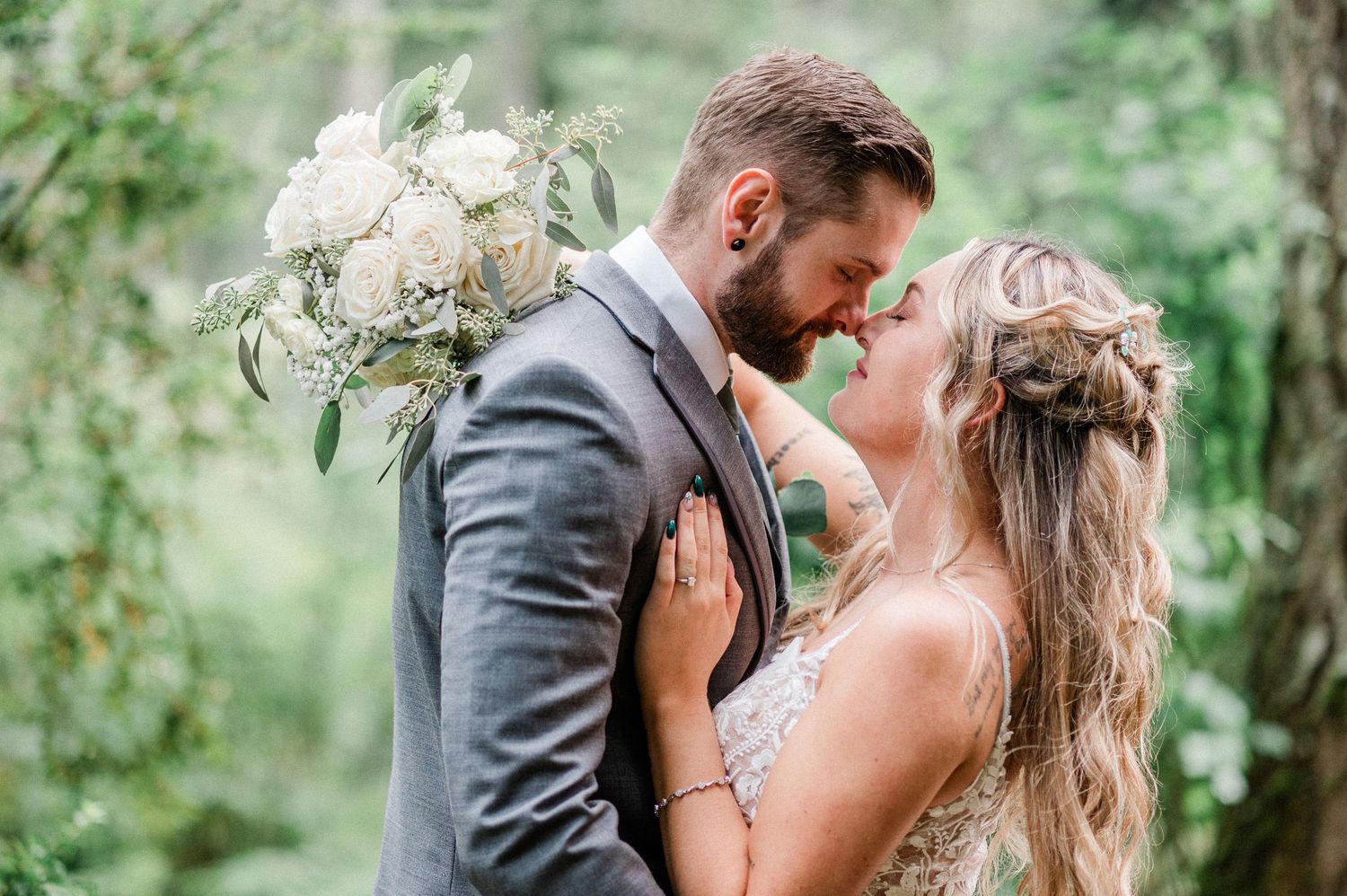 A bride and groom share a romantic moment outdoors, surrounded by lush greenery, as she holds a white floral bouquet.