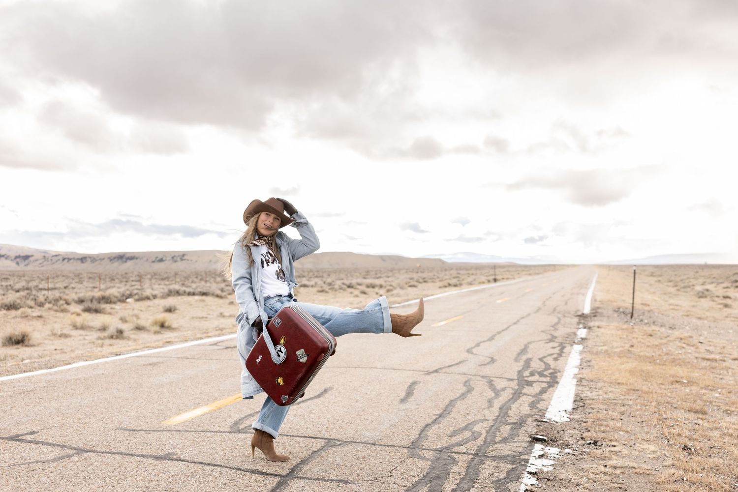 Woman in cowboy hat hitchhiking on desert highway with vintage suitcase, camera, and cardboard sign.