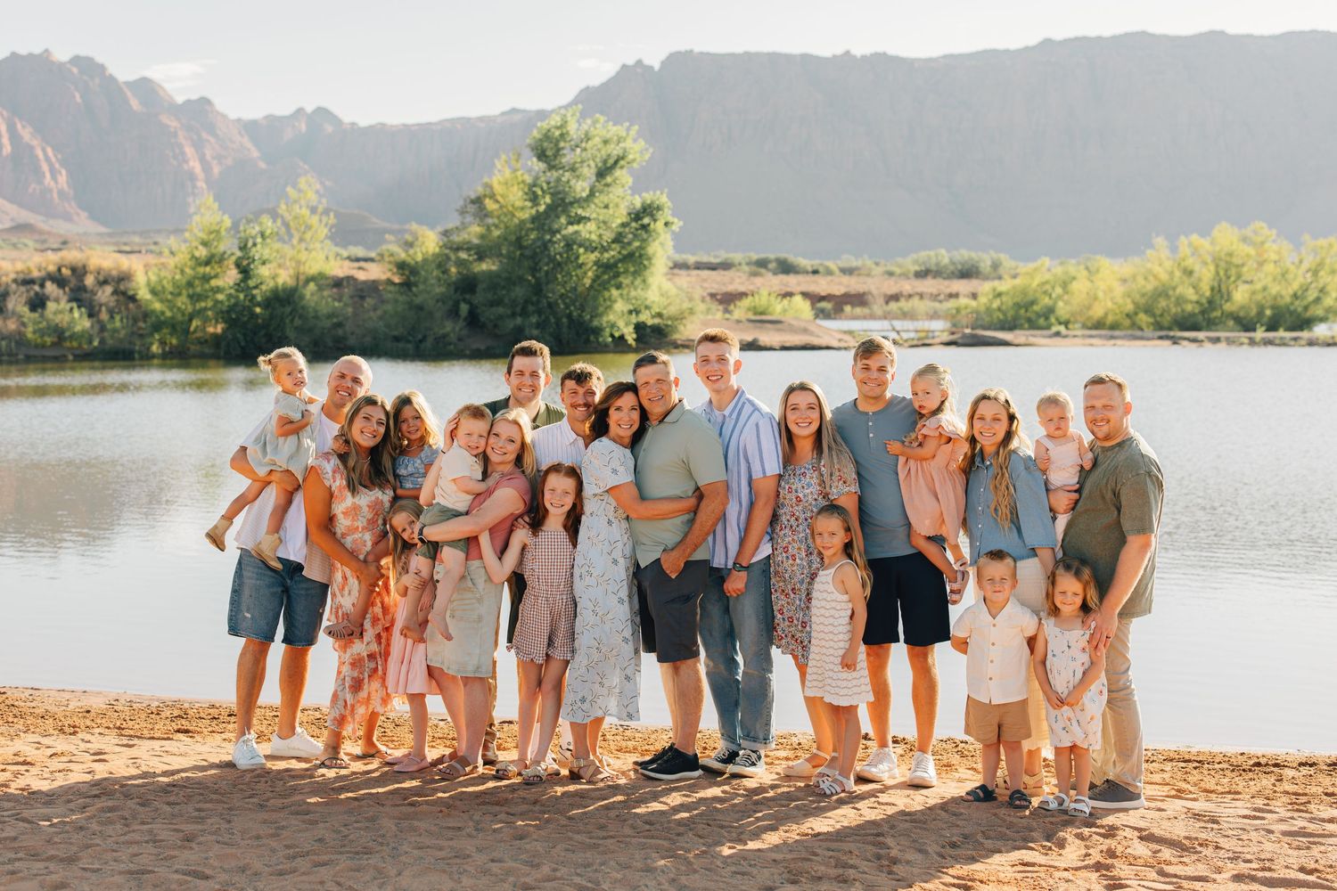 Large family group photo outdoors by a lake with mountain backdrop on a sunny day.