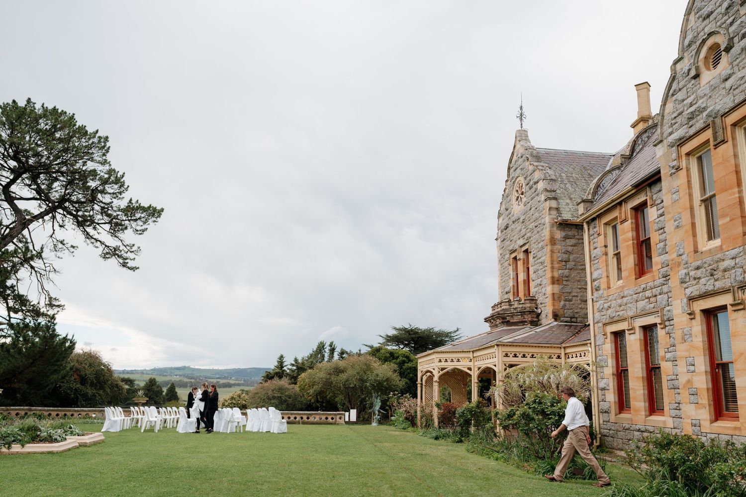 Elegant stone manor wedding venue with manicured lawn, white chairs, and guests gathering under cloudy skies.