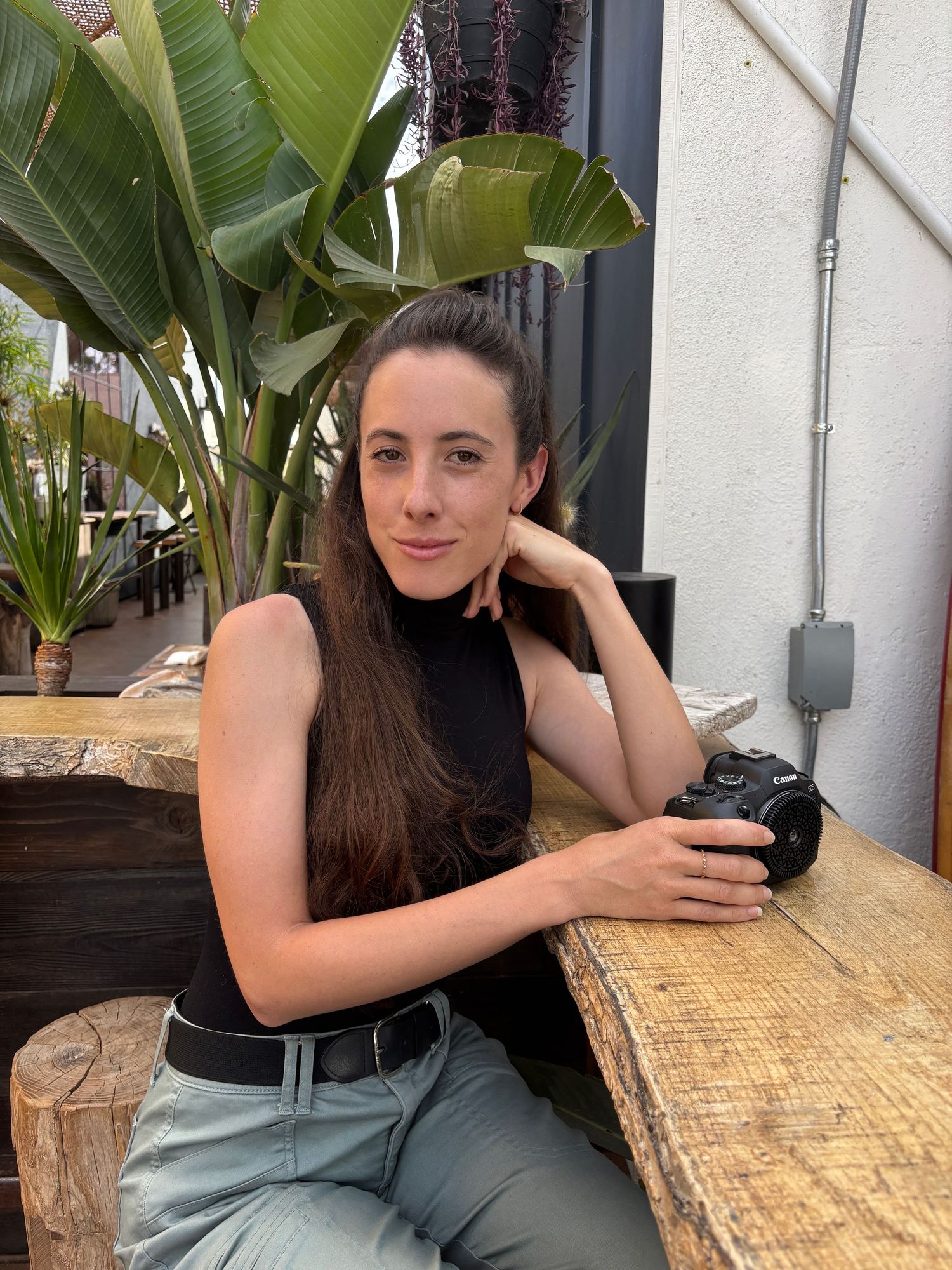 Young woman with long brown hair holding a camera, seated at a wooden table with tropical plants in the background.