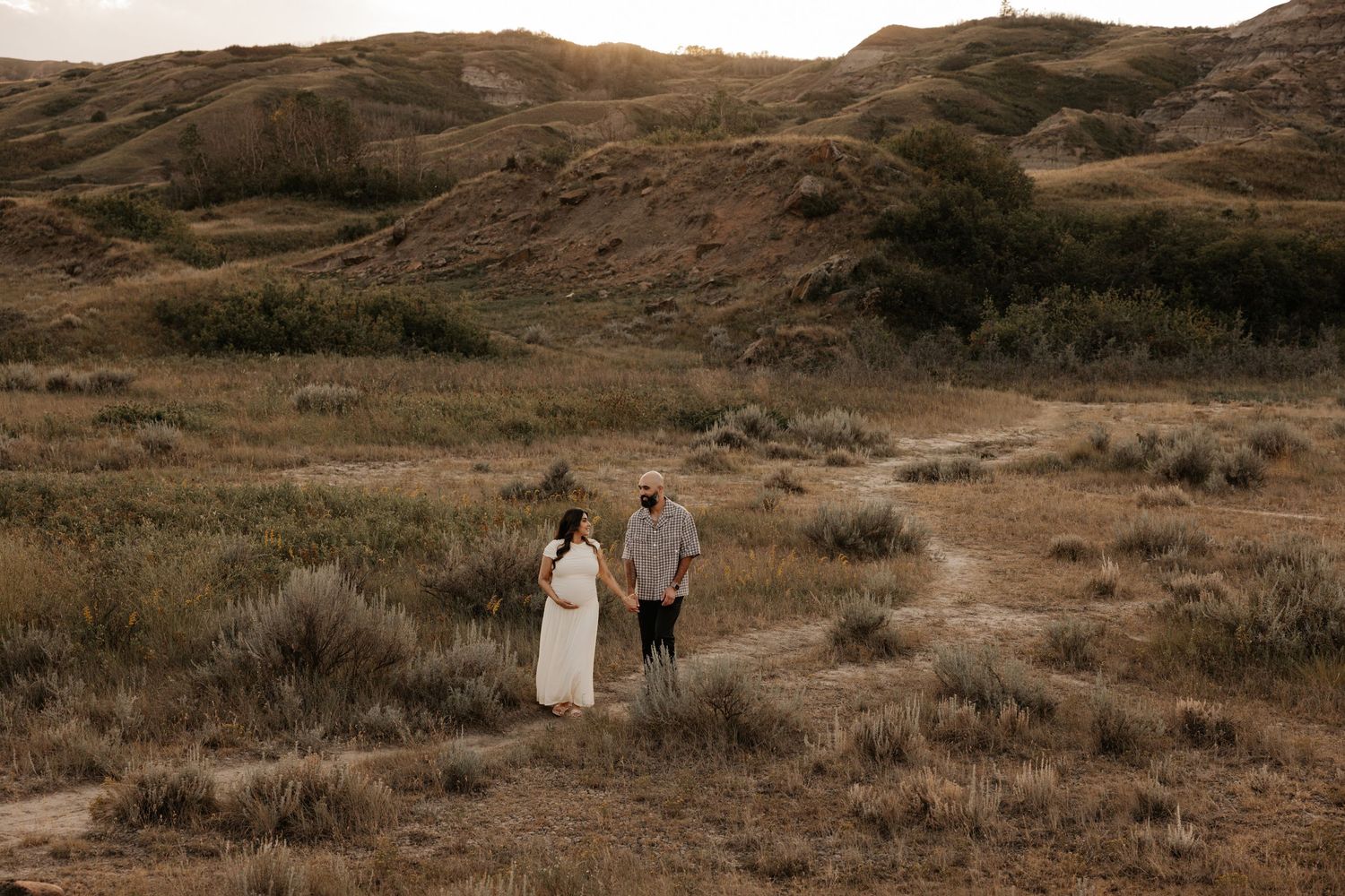Couple holding hands in vast arid badlands landscape, woman in white dress, golden hour light.