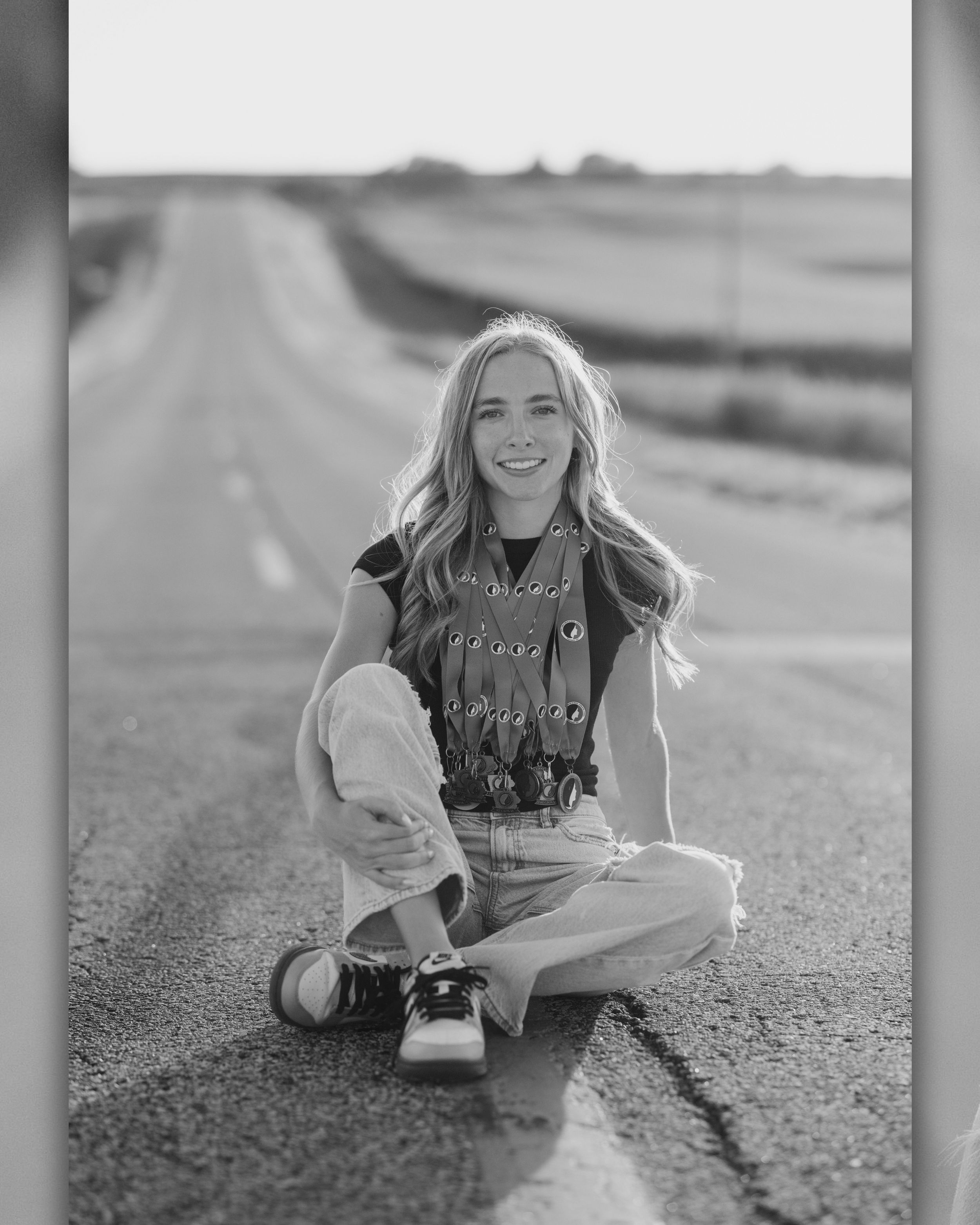 Black and white photo of a smiling young woman sitting cross-legged on a long straight road stretching into the horizon.