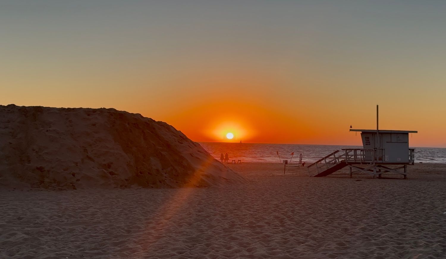Golden sunset over sandy beach dunes with lifeguard station silhouette against vibrant orange sky.