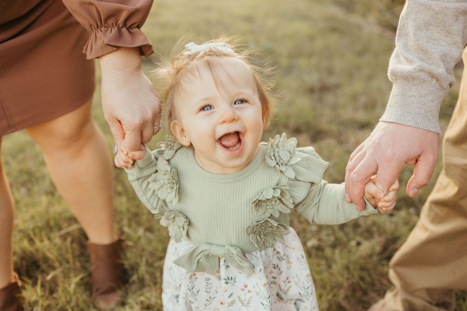 Smiling toddler in a sage green ruffled top holding hands with two adults while walking through a sunny grassy field.