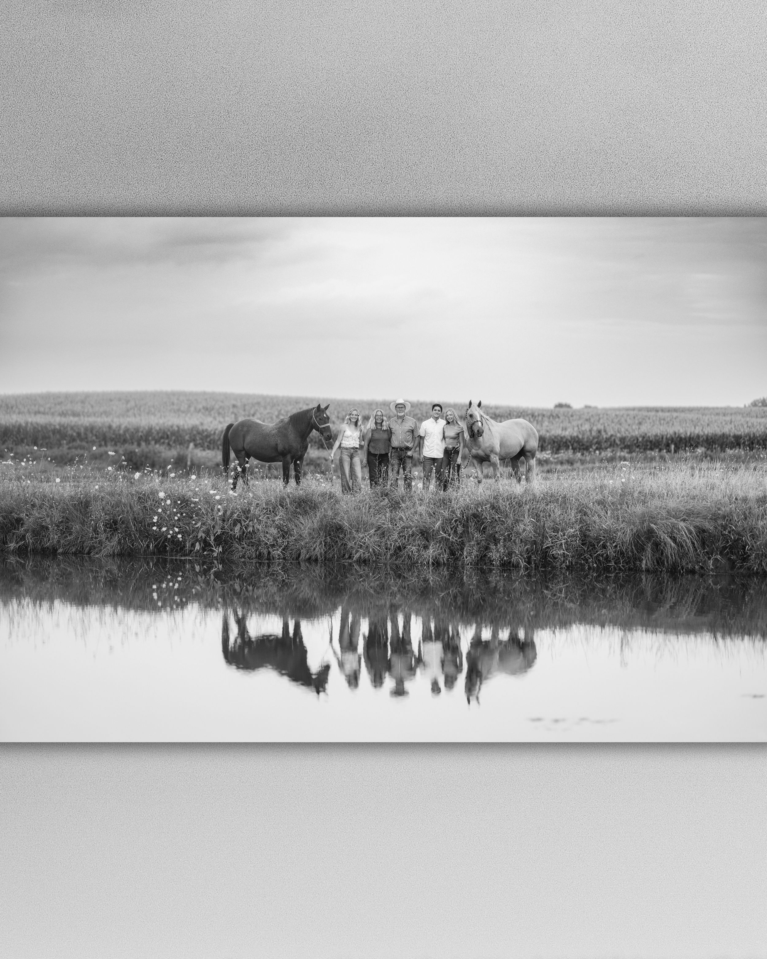 Black and white photo of a group of people standing between two horses beside a calm pond, with their reflections in the water. JADE MADE