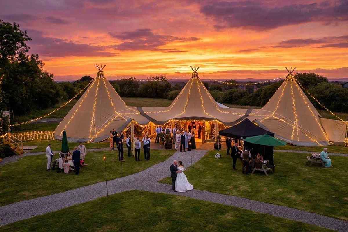 Bride in a white gown walks toward a glowing tipi wedding reception as guests gather on the lawn under a vivid orange and purple sunset sky.