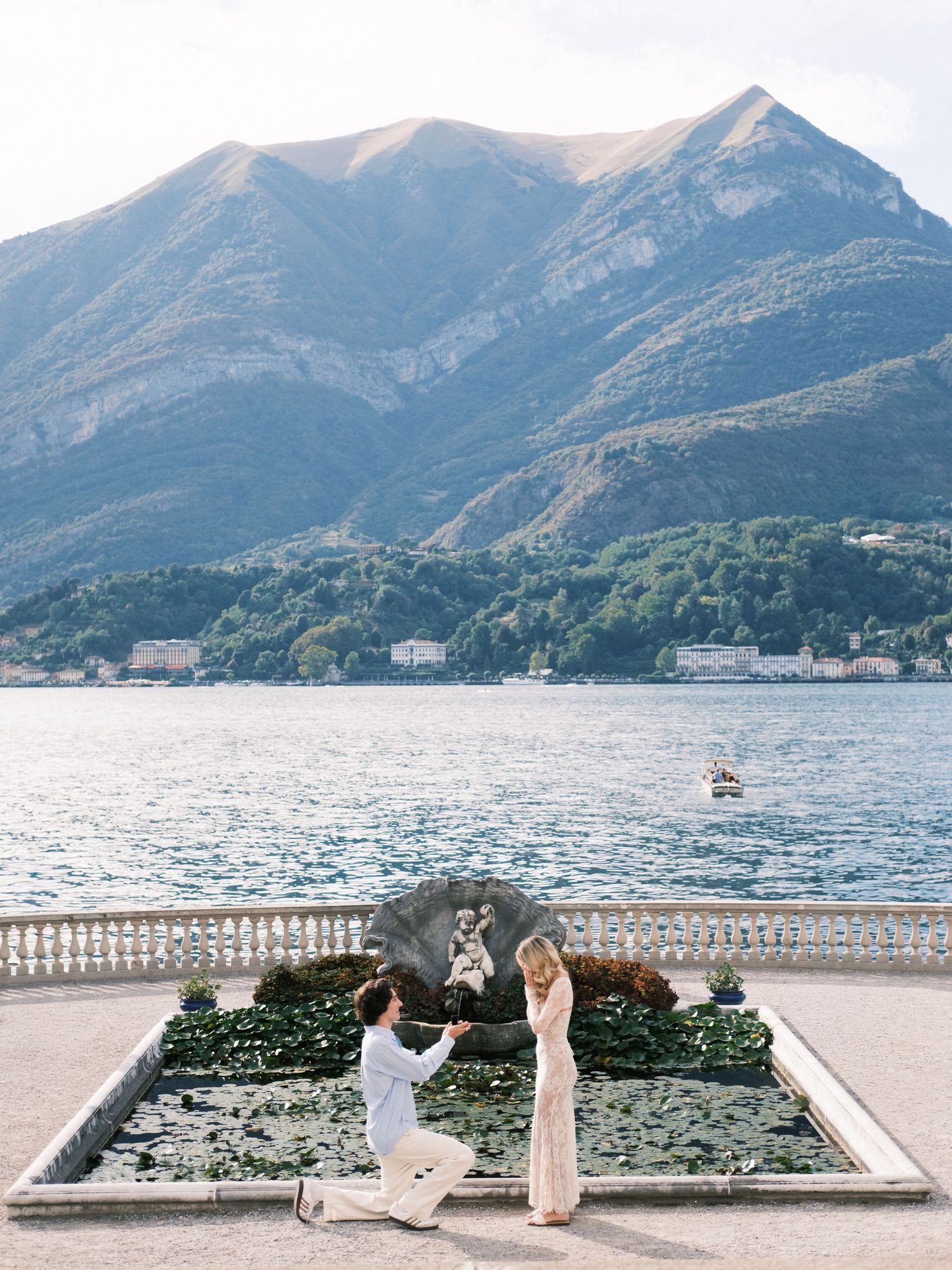 Man proposes to woman on one knee by Lake Como, Italy, with stunning mountain backdrop and ornate garden.