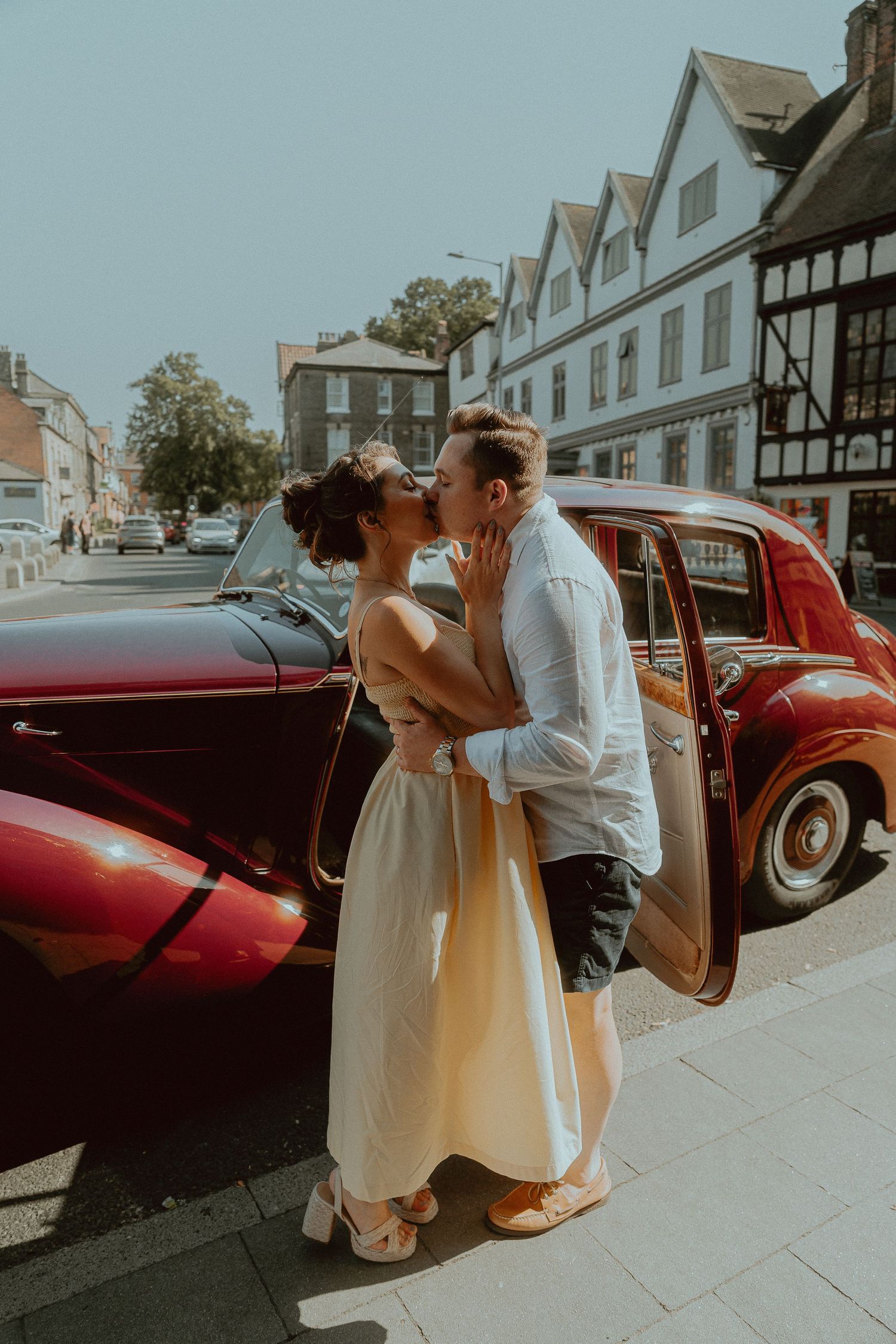 Couple sharing a romantic kiss beside a vintage red car on a sunny street in Norwich Norfolk, bride in elegant white skirt.