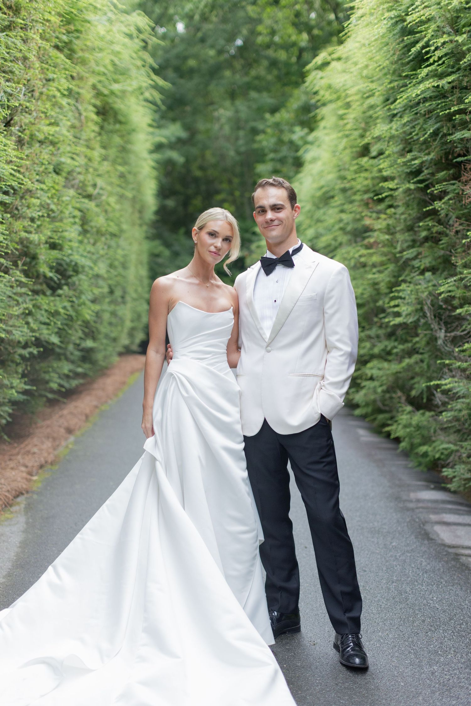 A couple in formal wedding attire poses together outdoors at sunset against a scenic backdrop.