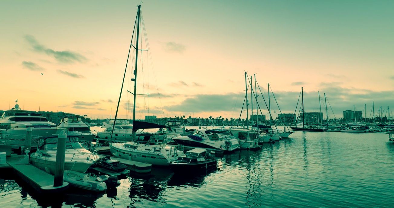 Sailboats and yachts docked at a marina during golden sunset with calm reflective water and colorful sky.