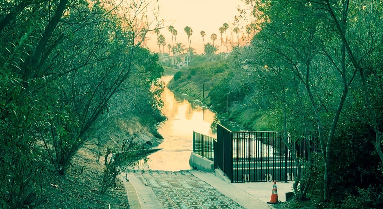 A serene canal winds through lush green trees at sunset, with a metal gate and orange traffic cone in foreground.