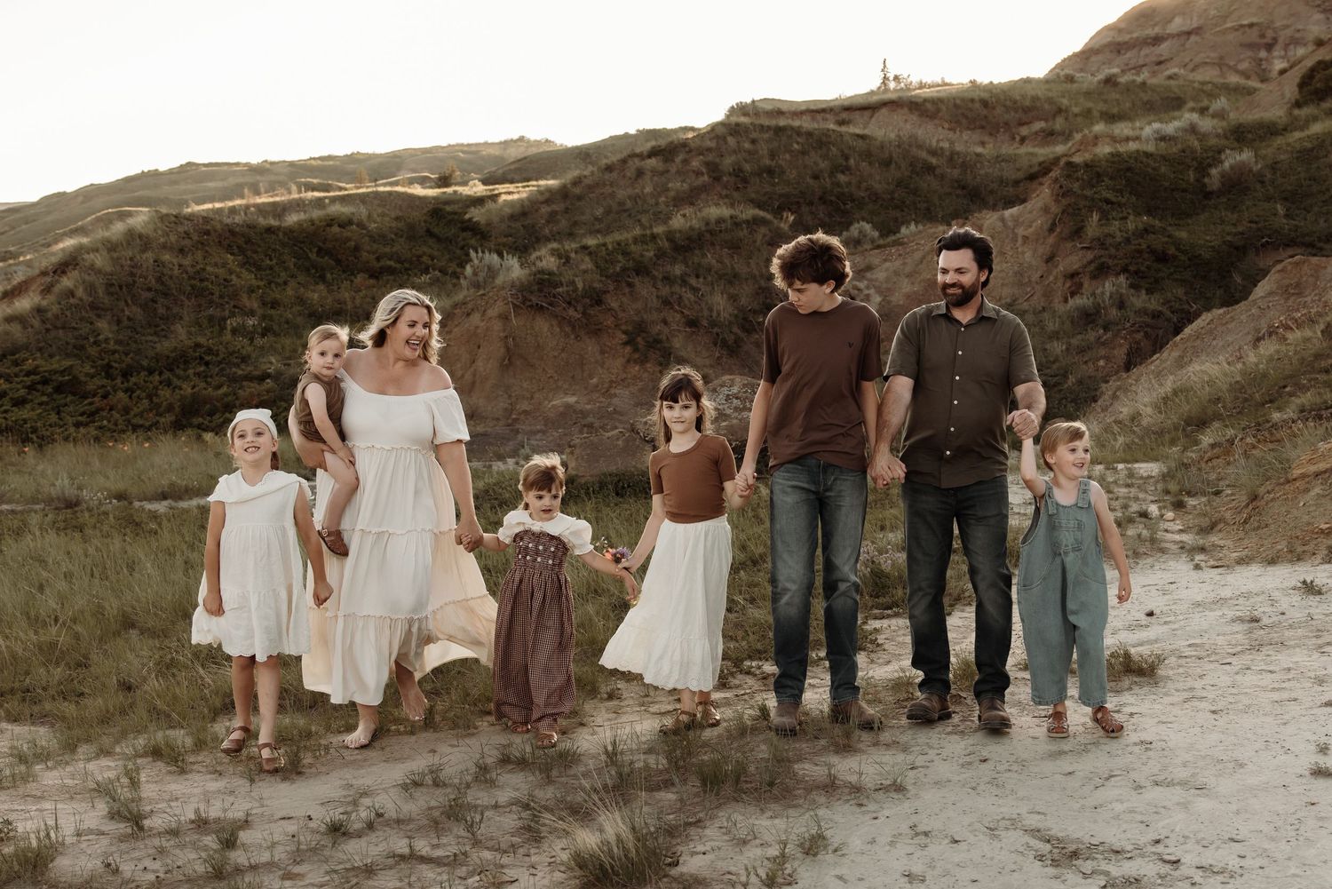 Family of seven posing outdoors on sandy terrain with green hills, wearing coordinated earth-tone outfits.