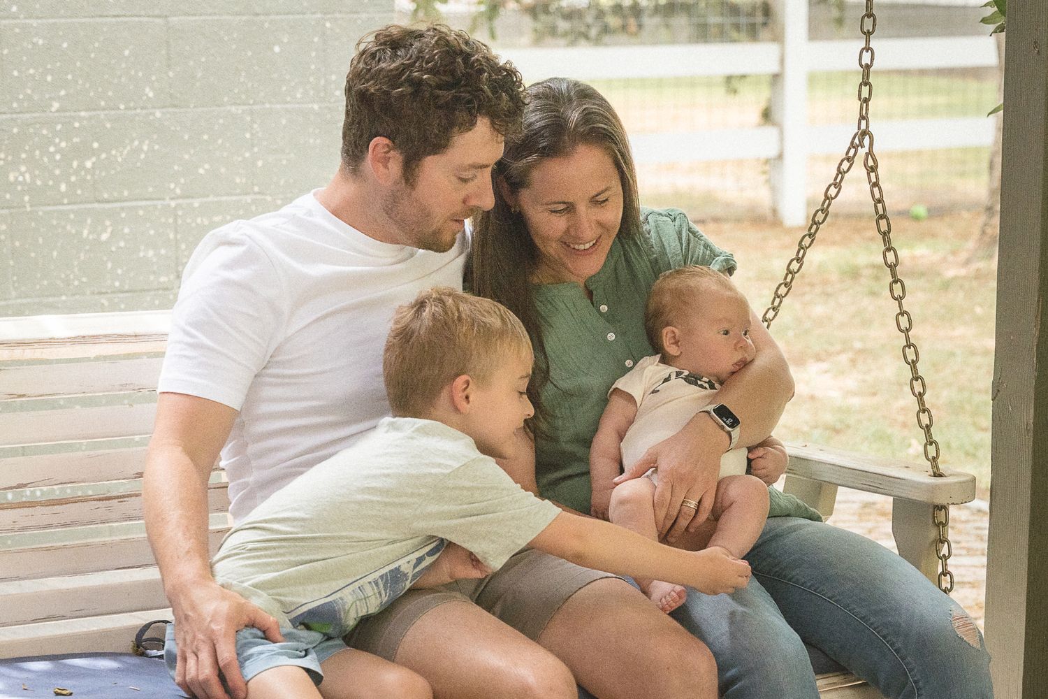 Happy family of four sits on a porch swing, parents smiling at newborn baby while young boy looks on.