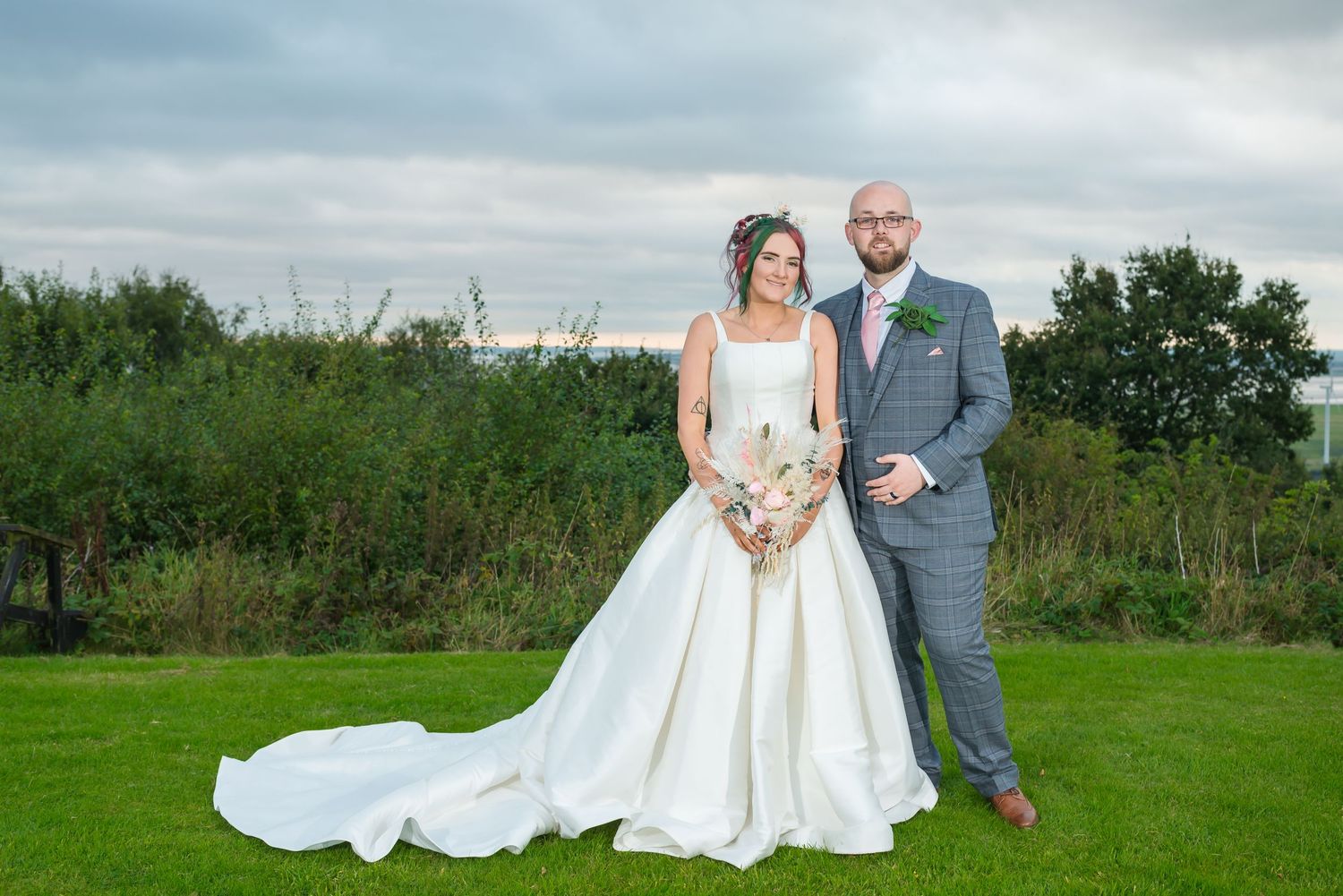 Bride in white ball gown and groom in grey suit pose together on green lawn at outdoor wedding venue.