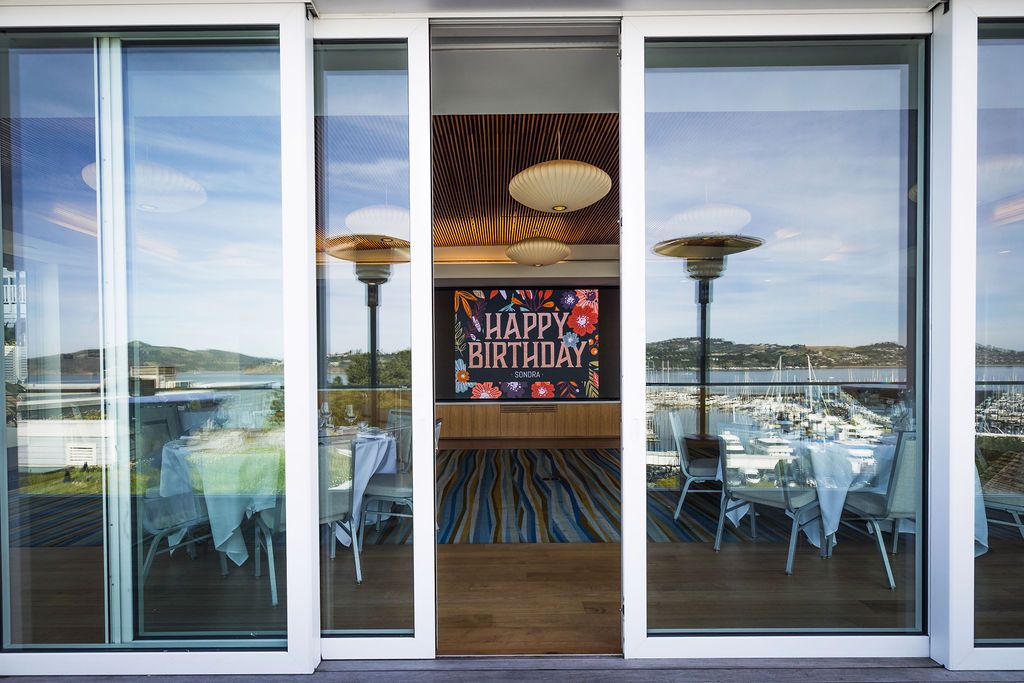 Indoor birthday party venue with sliding glass doors, patio heaters, dining tables, and a Happy Birthday banner overlooking a marina.