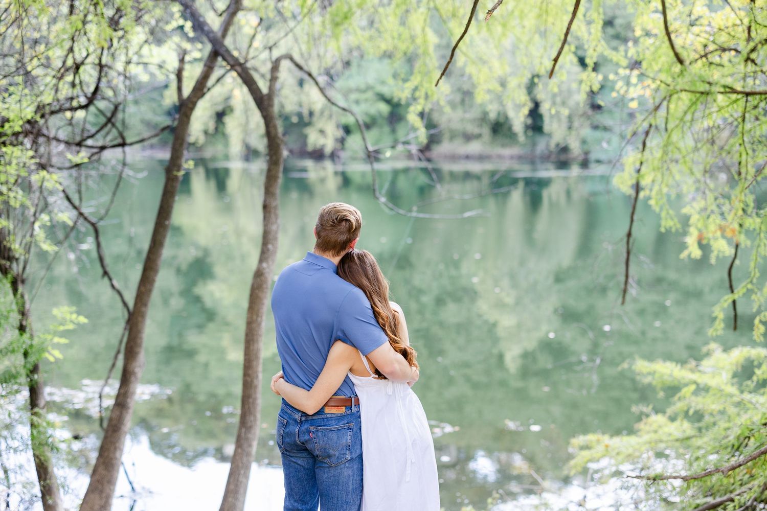 Couple embracing by a serene green lake surrounded by lush trees in a romantic outdoor setting.