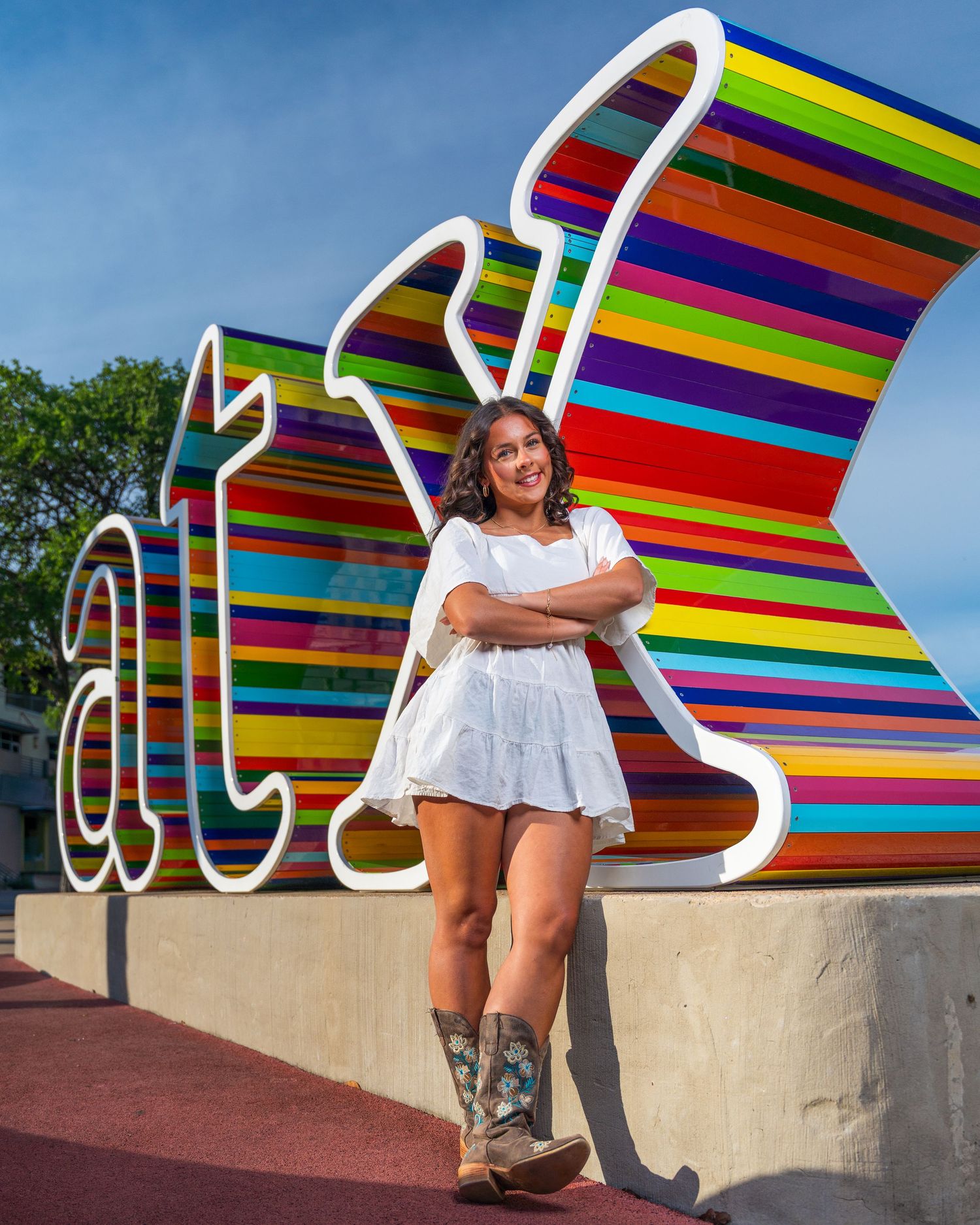 Young woman in white dress and cowboy boots poses confidently in front of colorful striped ATX sign in Austin, Texas.