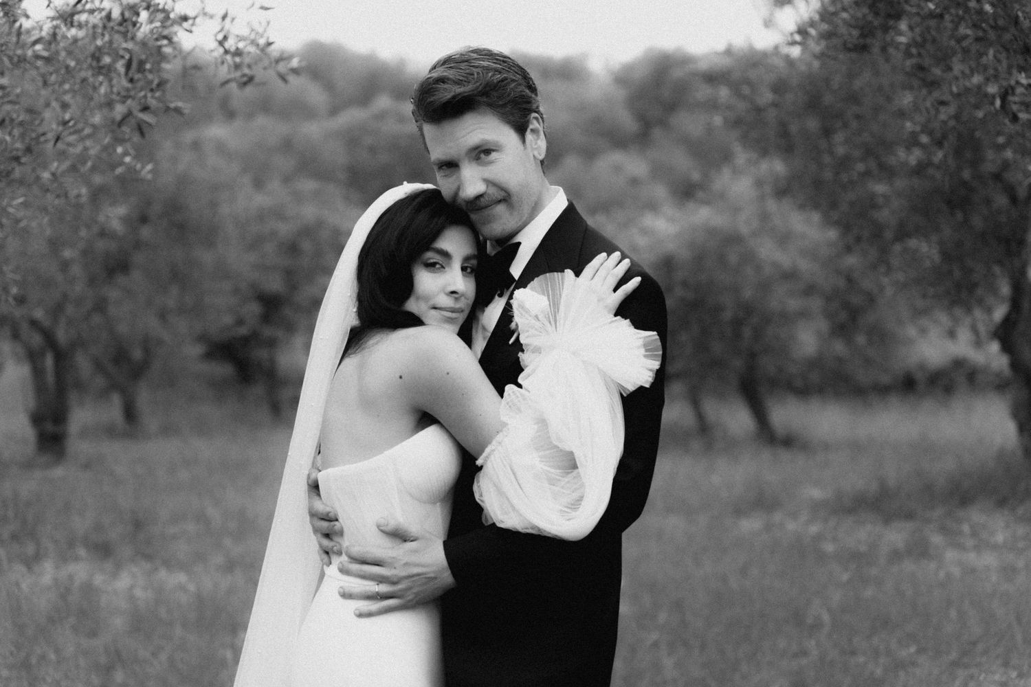 Black and white wedding photo of a couple embracing outdoors among trees, bride in strapless gown, groom in tuxedo.