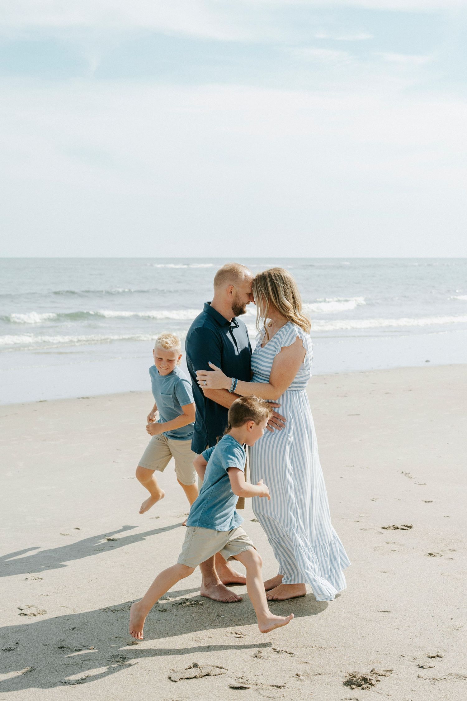 Family of four enjoying a sunny beach day, two boys running while parents embrace near ocean waves.