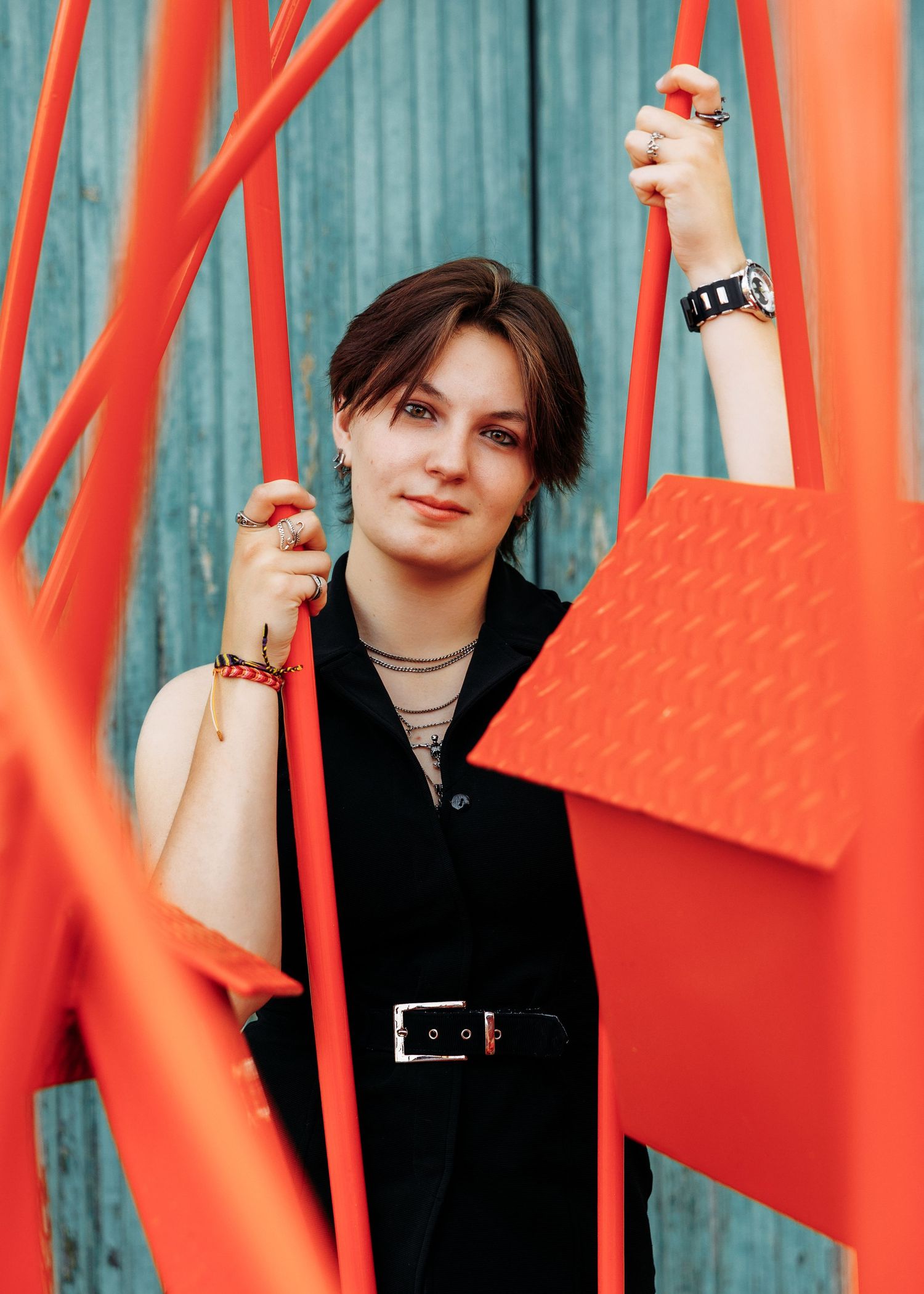 Young woman in black outfit poses among vibrant red metal structures against a teal wooden wall.