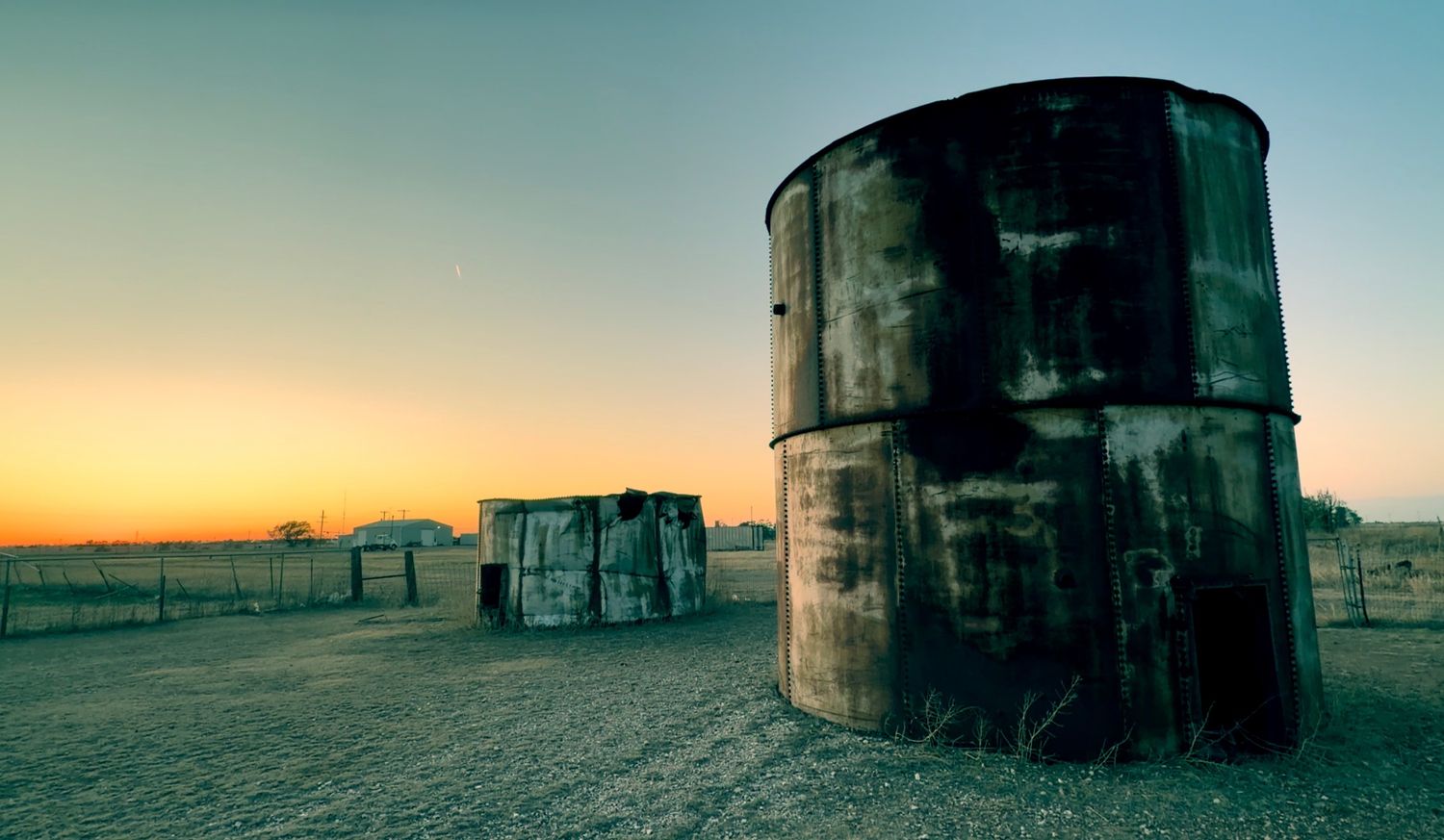 Rusty industrial oil drums stand in a barren field at sunset, creating a dramatic abandoned landscape scene.
