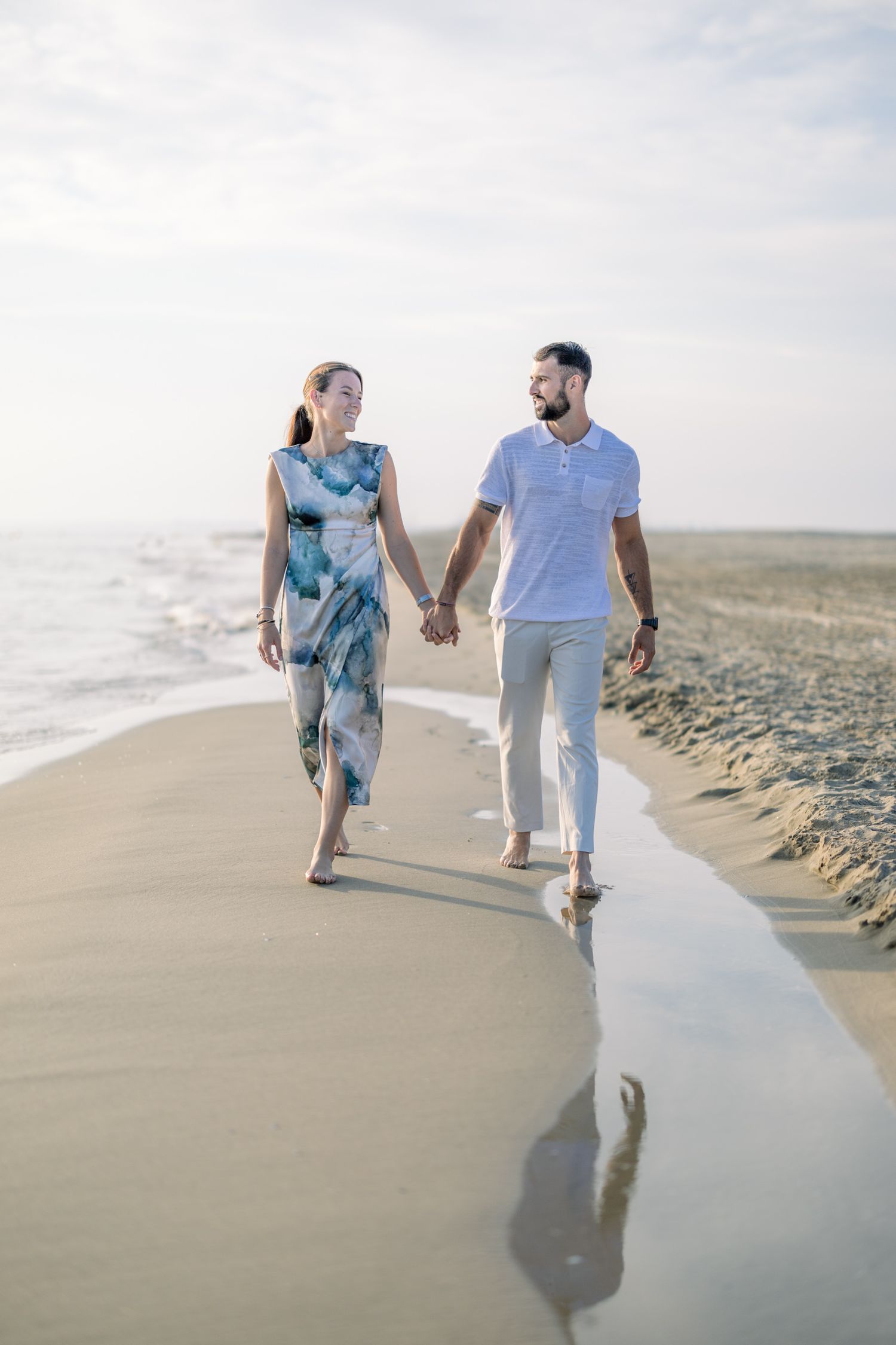 A couple walks hand in hand along a serene beach shoreline with waves gently rolling in on a cloudy day.