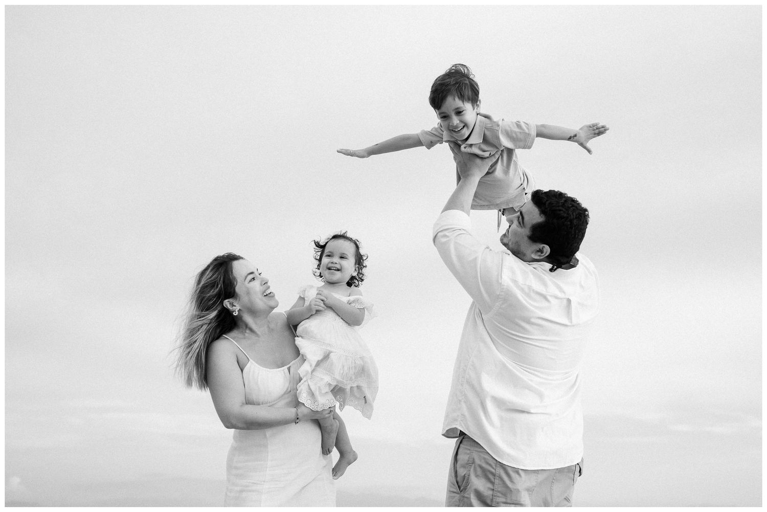 A family plays together on a beach, lifting a child in the air while wearing white clothing in black and white photography.