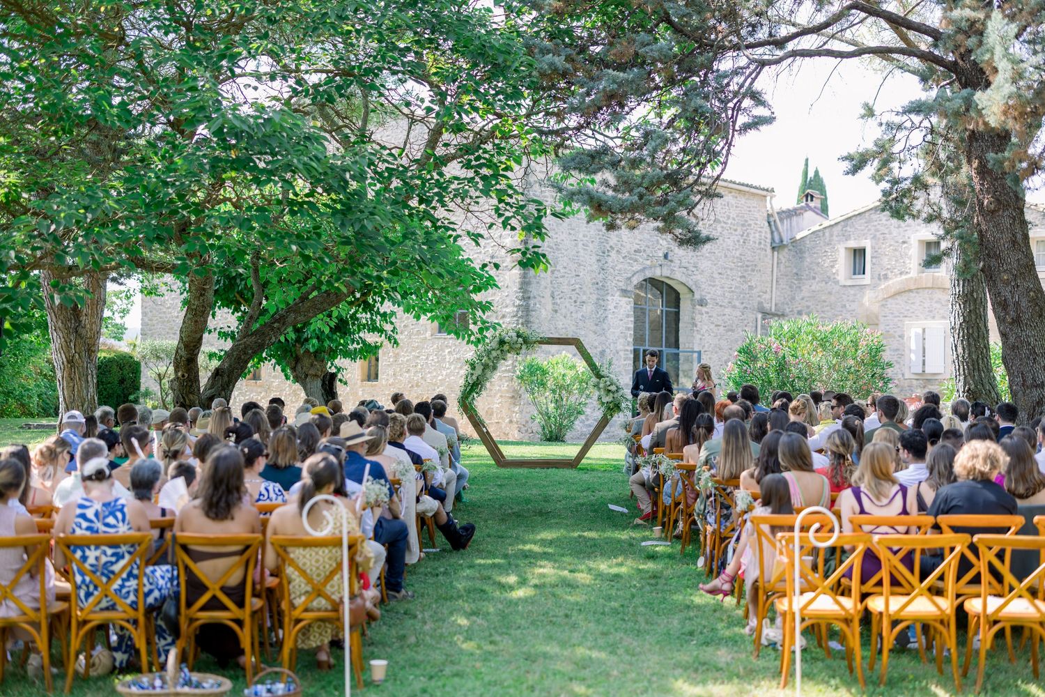 Outdoor wedding ceremony guests seated on wooden chairs beneath lush green trees on a sunny day.