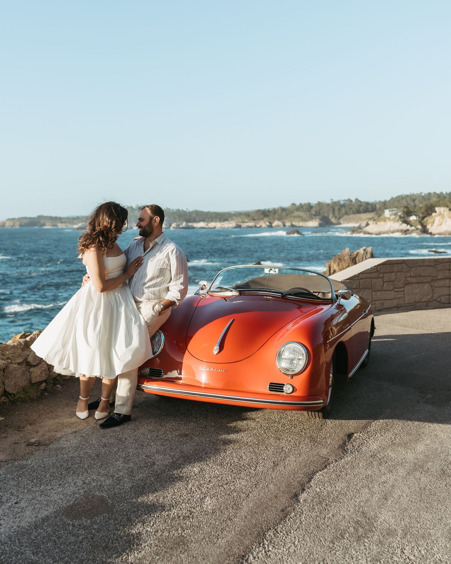 Couple embracing by the ocean next to a classic dark blue convertible car on a scenic coastal road.