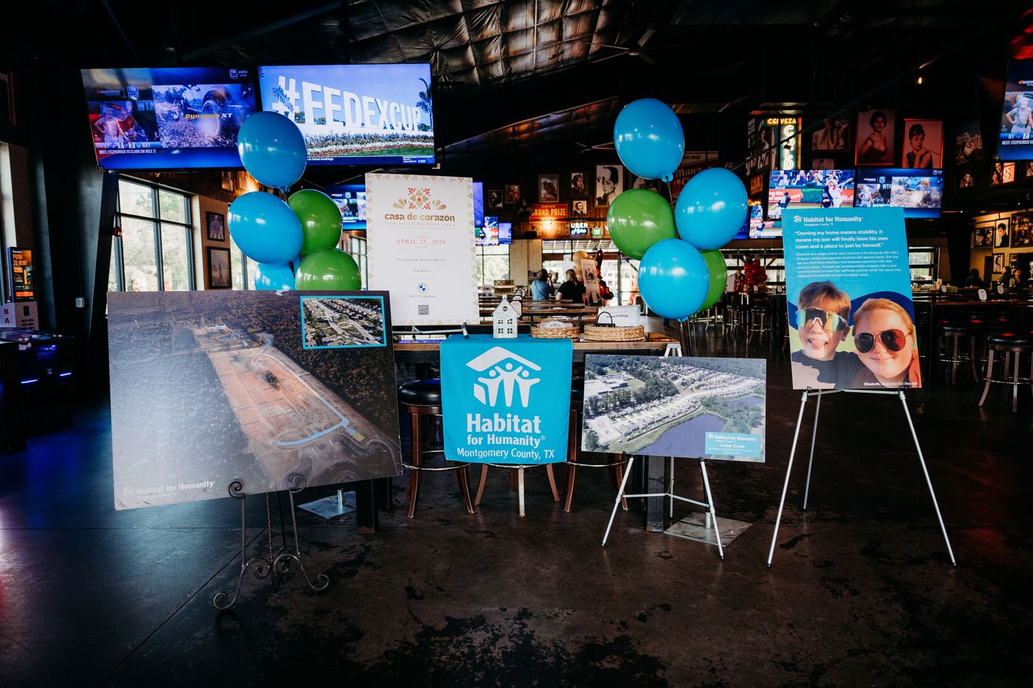 Event setup at a dark venue with blue and green balloons, signage, and tables arranged for a gathering.