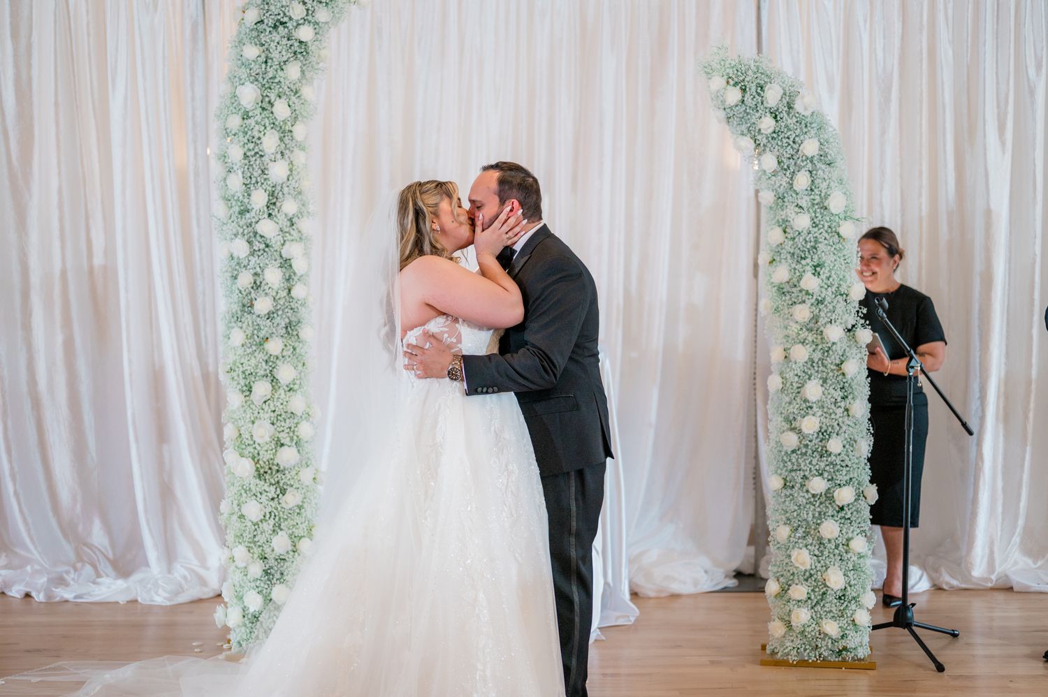 A couple shares a romantic kiss during their wedding ceremony in front of a white curtain backdrop with floral decor.