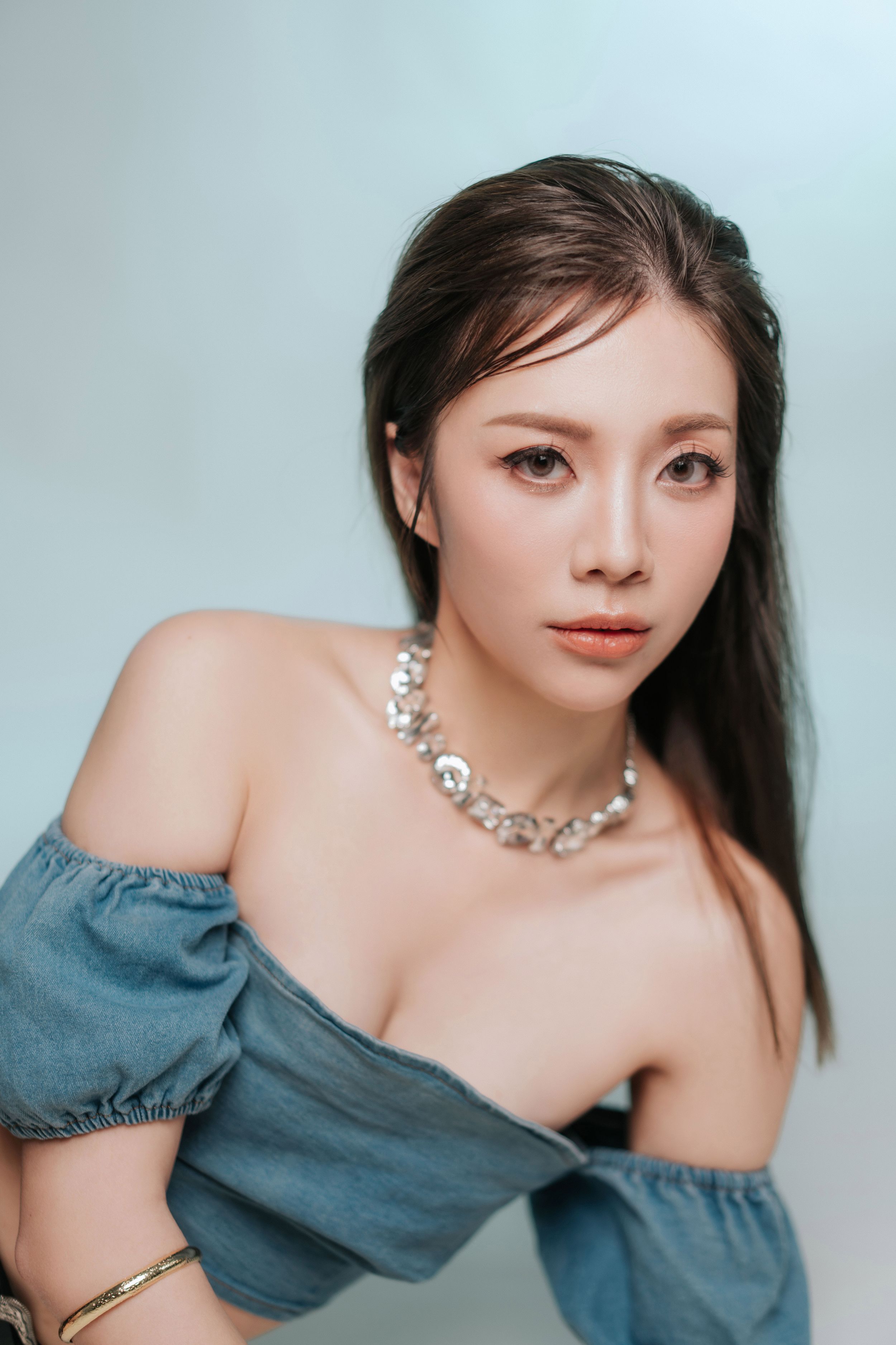 Young Asian woman in off-shoulder blue top and silver chain necklace posing elegantly against light background.