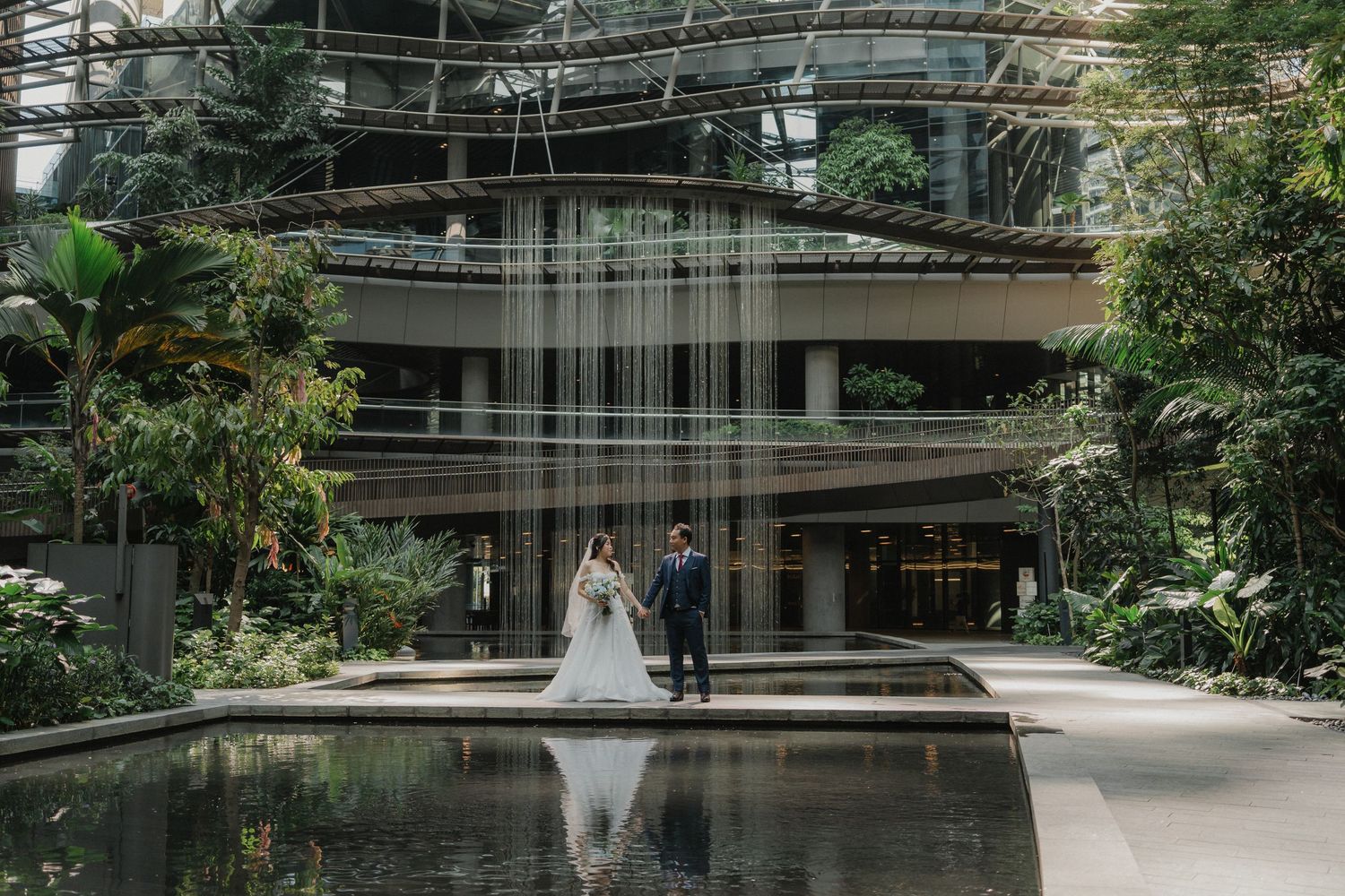 Two people stand by a reflective pond surrounded by lush greenery and modern architecture with cascading water features.
