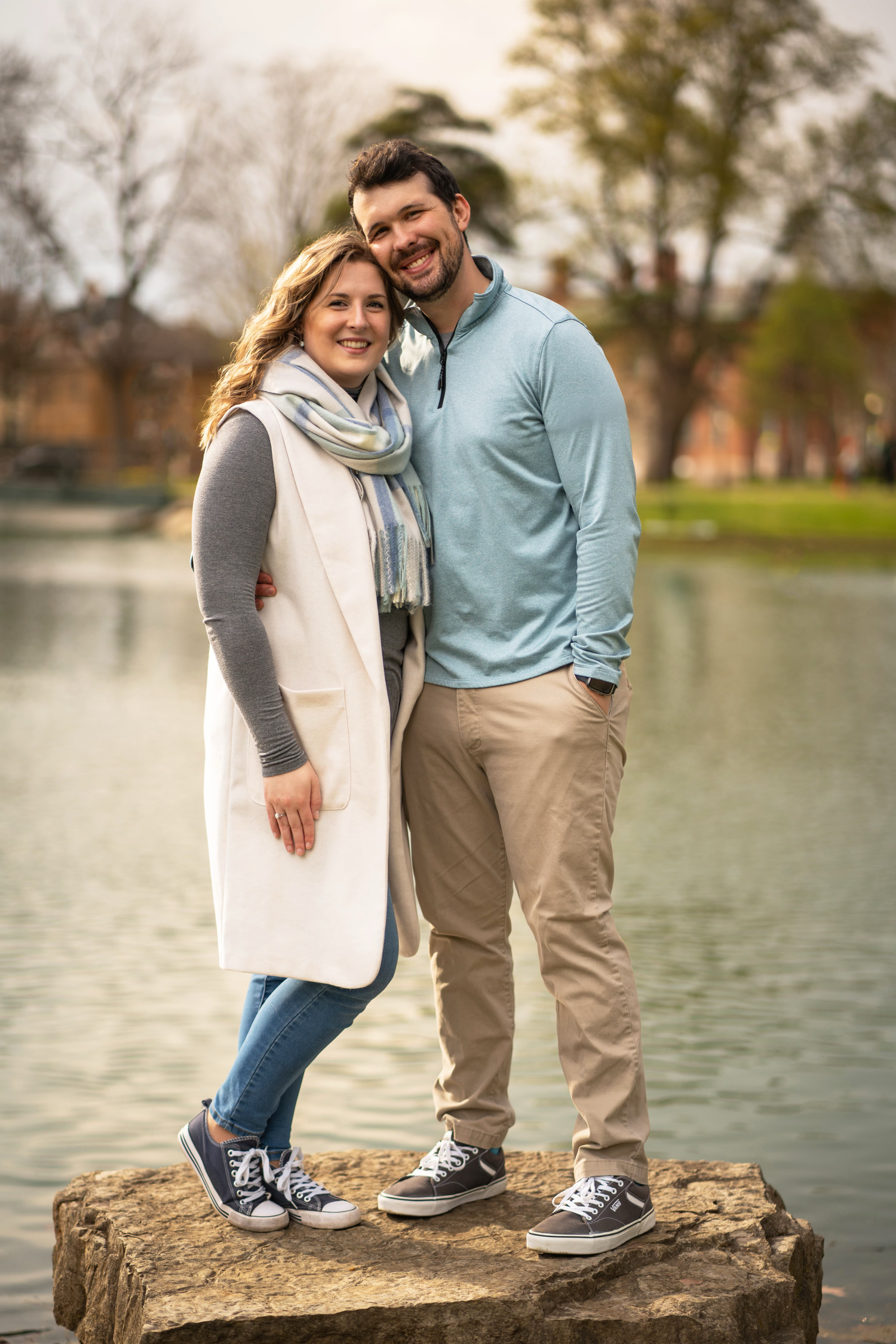 A couple wearing casual clothing poses playfully near a lake, showing only their feet and legs on a rock.