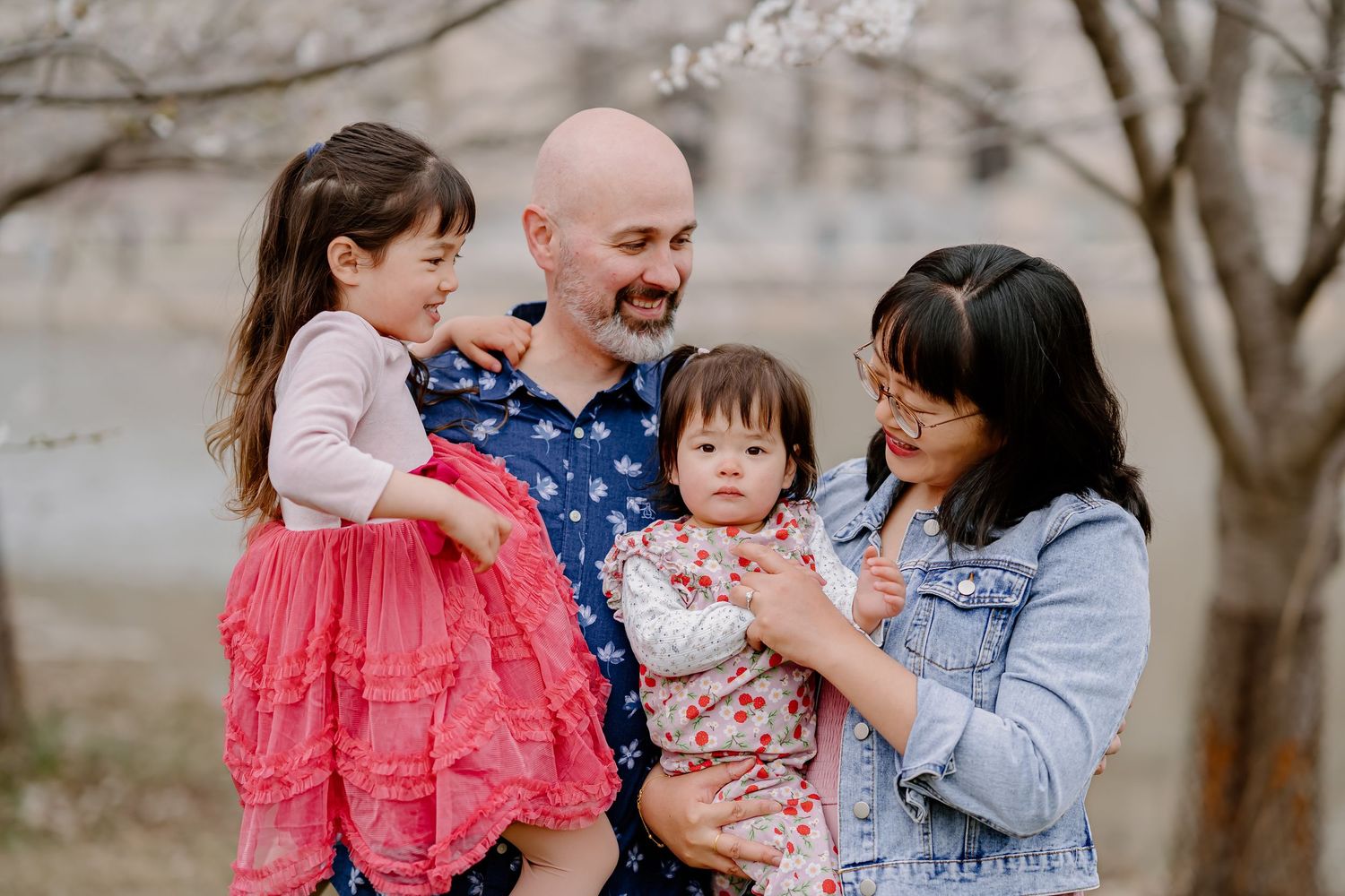 Family of four posing under blooming cherry blossom trees near a lake in spring.