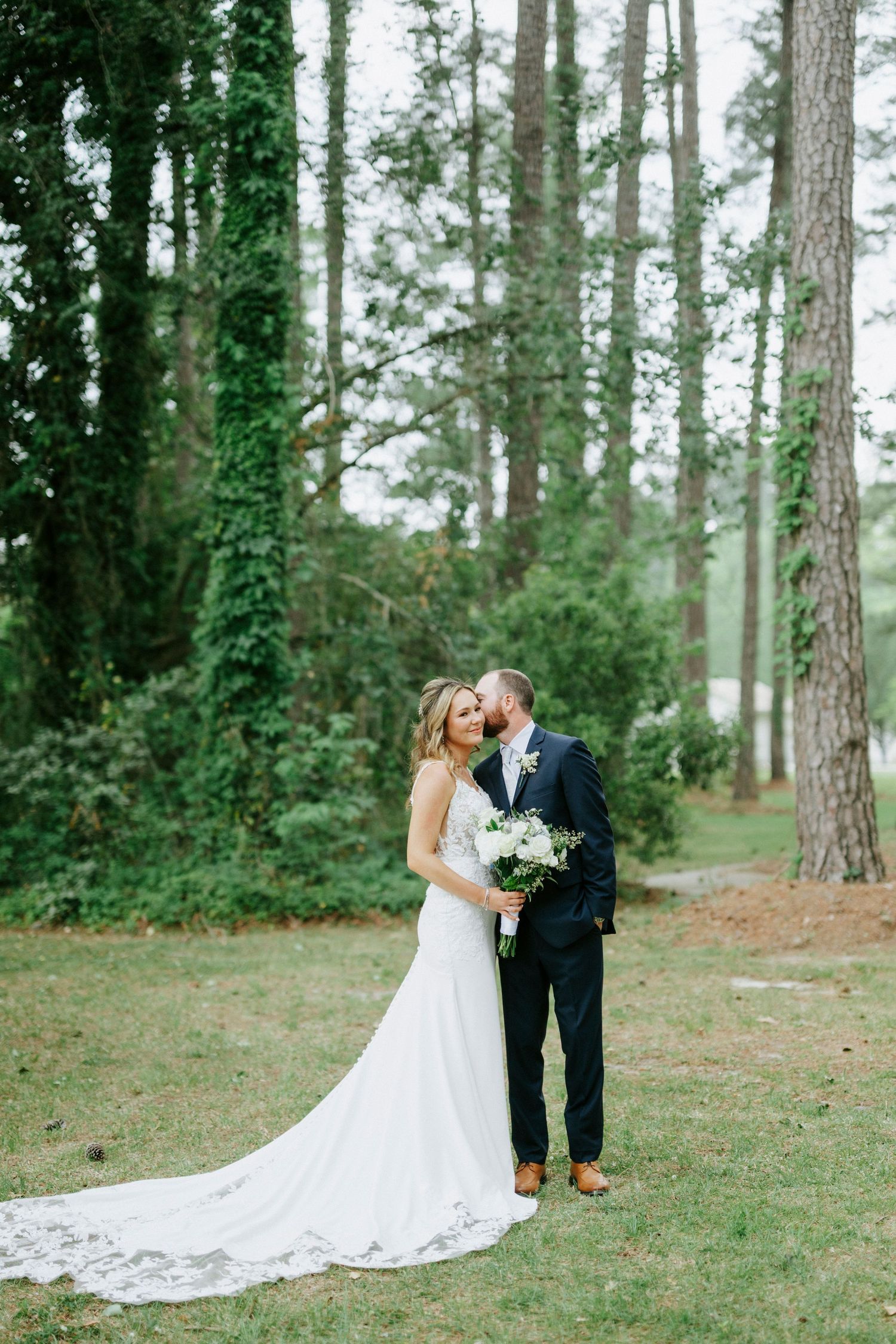 Bride and groom share a romantic moment in a lush green forest, bride holding a white bouquet in an elegant gown.