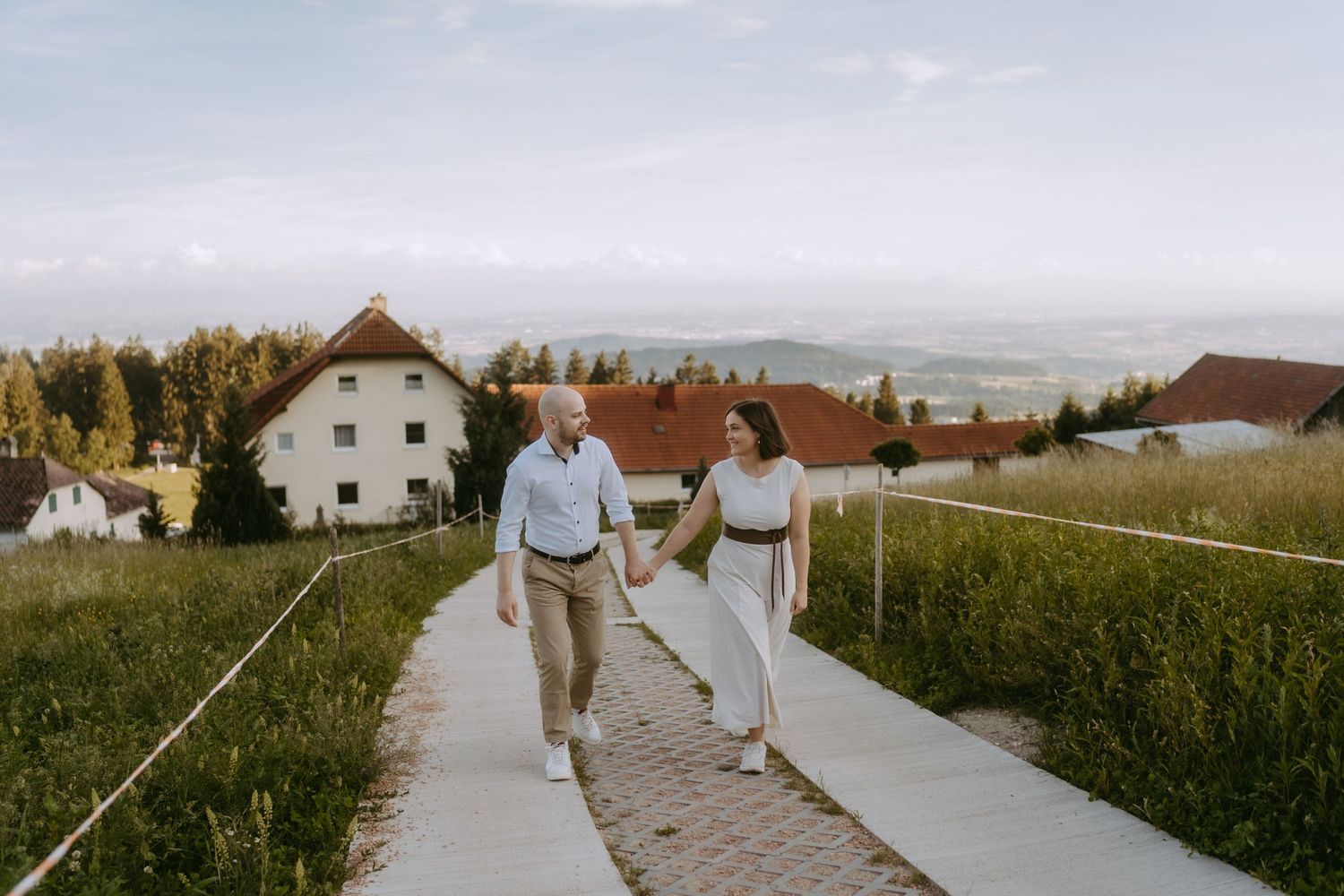 Couple holding hands walking on a cobblestone path with green hills and white buildings in the background.