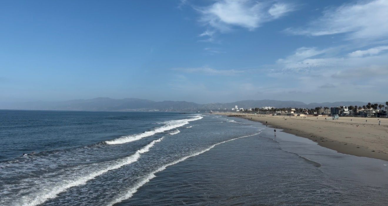Aerial view of a sandy beach with rolling waves, blue sky, and a coastal cityscape stretching along the shoreline.